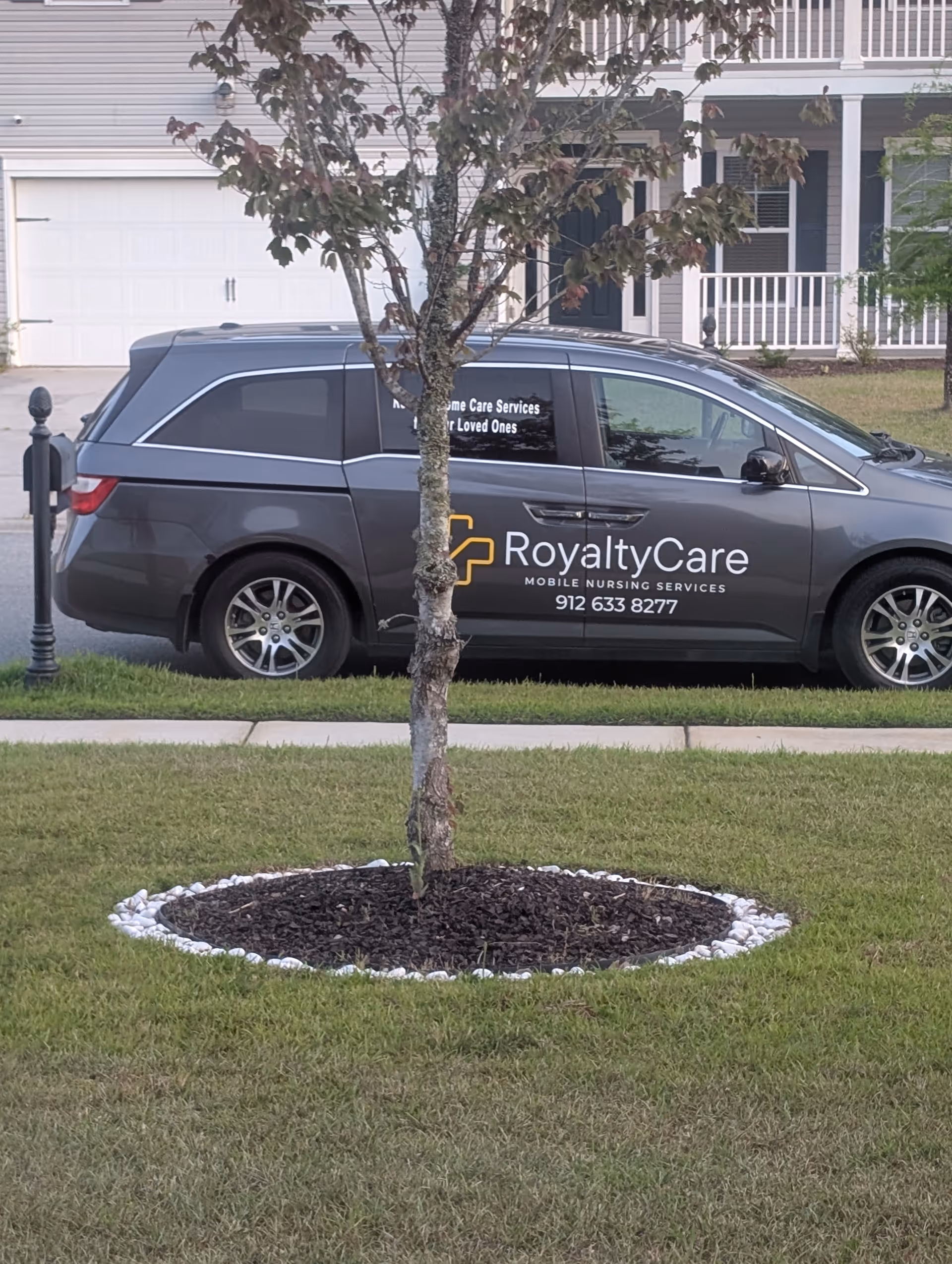 Gray RoyaltyCare mobile nursing van parked in front of a residential home with a tree