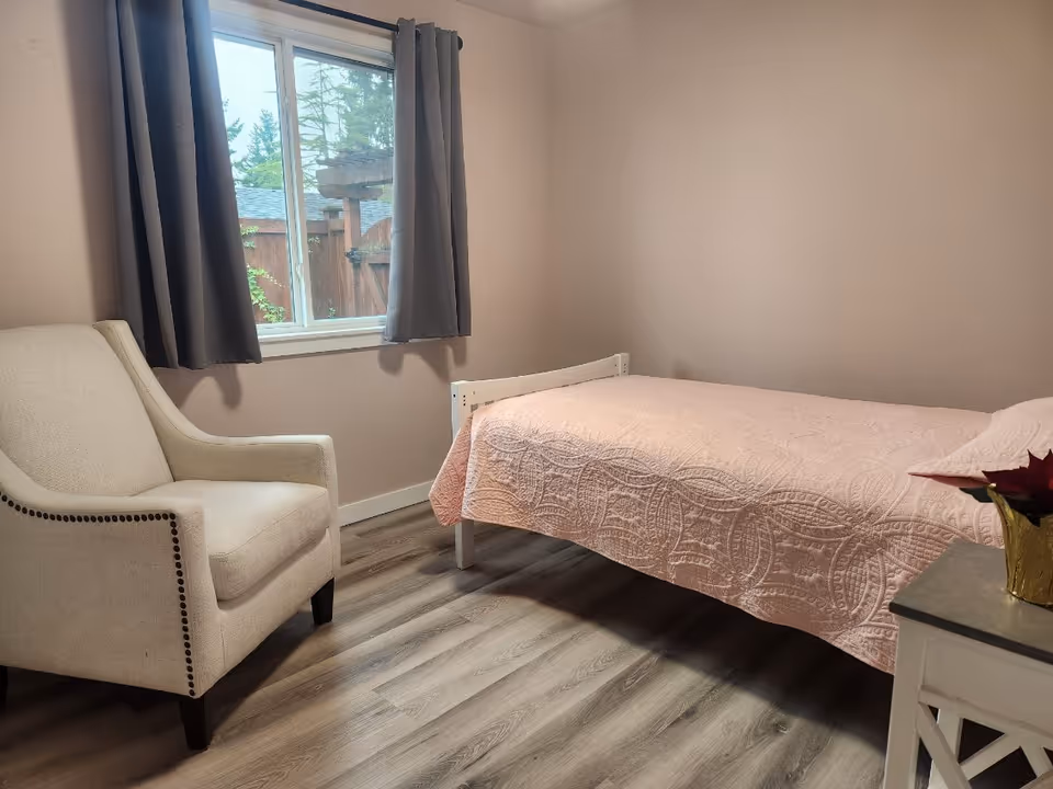 A cozy bedroom featuring a single bed with a pink quilt, an armchair, and a small table by the window.