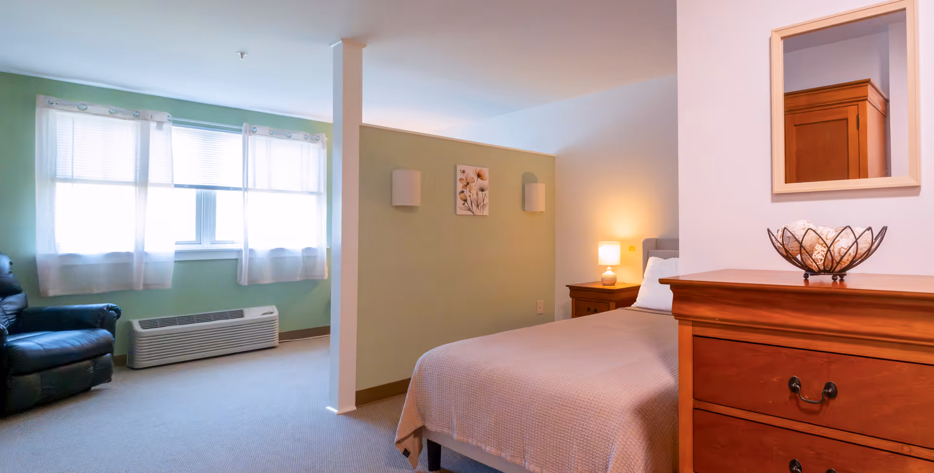 Well-lit assisted living bedroom with bed, dresser, nightstand, and sitting area.