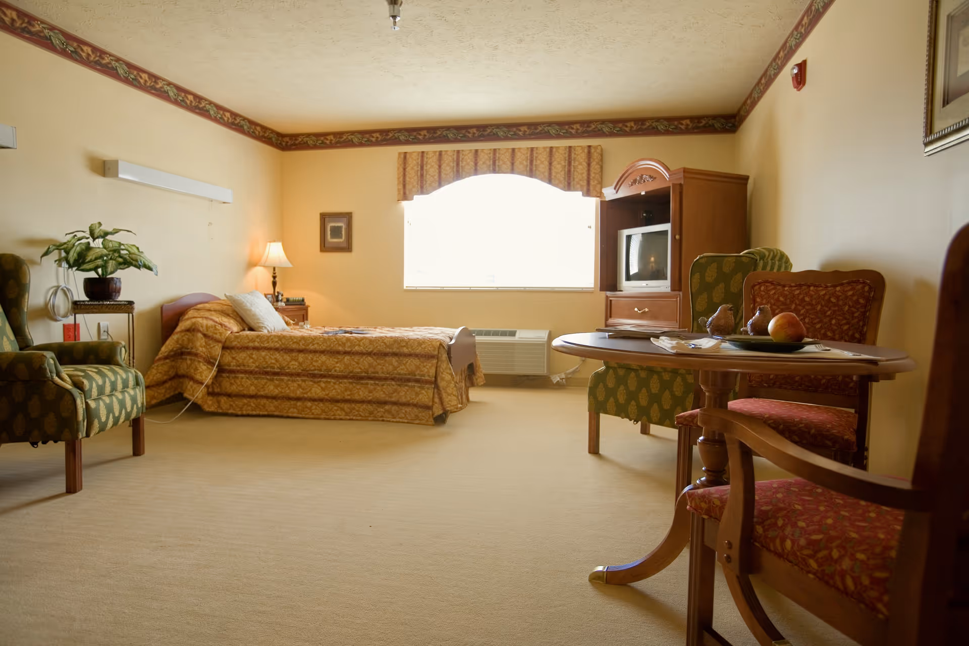 A cozy senior living bedroom with a single bed covered in a patterned bedspread, a wooden nightstand with a lamp, two upholstered armchairs, a round wooden table with fruit and decorative items, a wooden TV cabinet with a small television, and a large window with a valance letting in natural light.