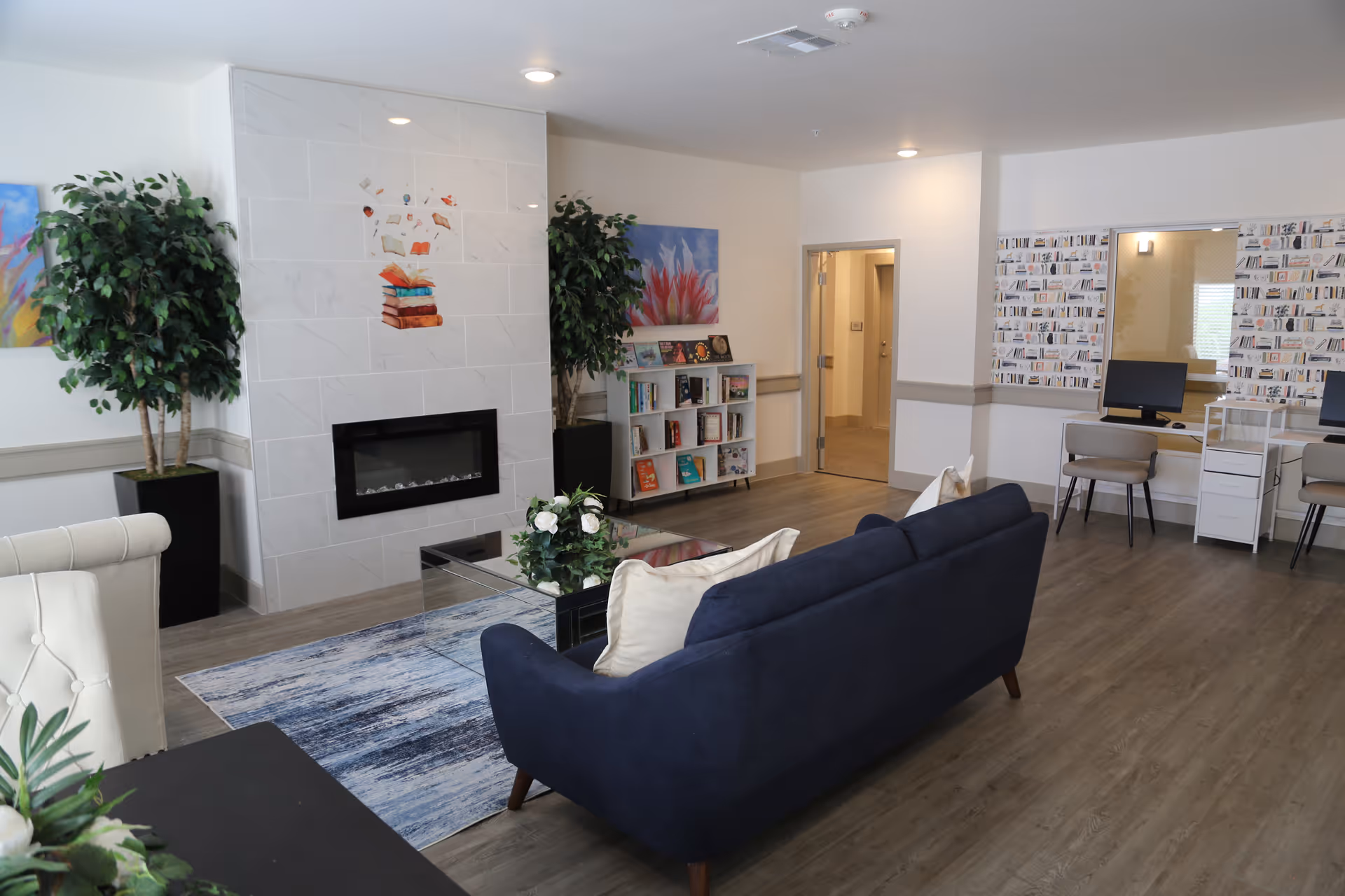 Modern senior living lounge with fireplace, navy sofa, rug, plants, and computer desks.