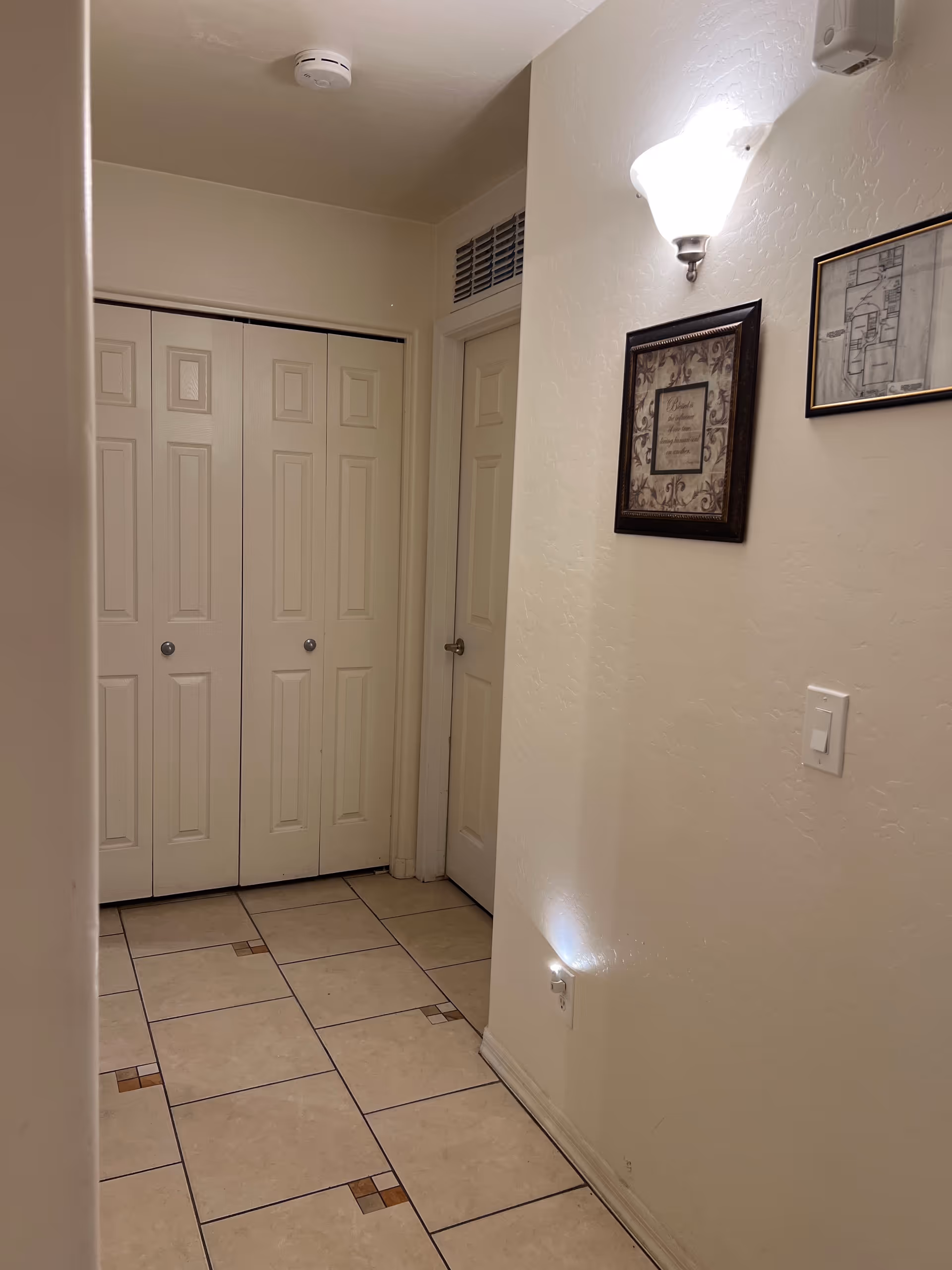 Narrow hallway with tile floor, closed closet doors, wall sconce, and framed art.