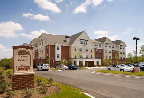 Four-story brick-and-siding apartment building with a front sign reading 'Cove Point' and a parking lot under a blue sky.