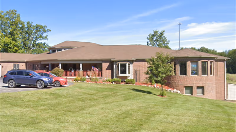 Single-story brick senior living facility with a manicured lawn, parked cars, and entrance porch.