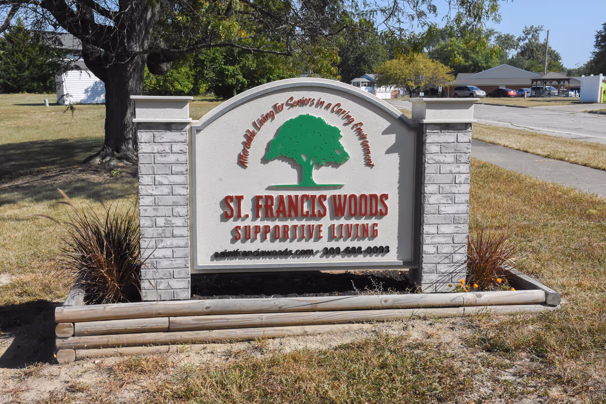 Outdoor sign at St. Francis Woods with a green tree logo, stone pillars, and surrounding landscaping.