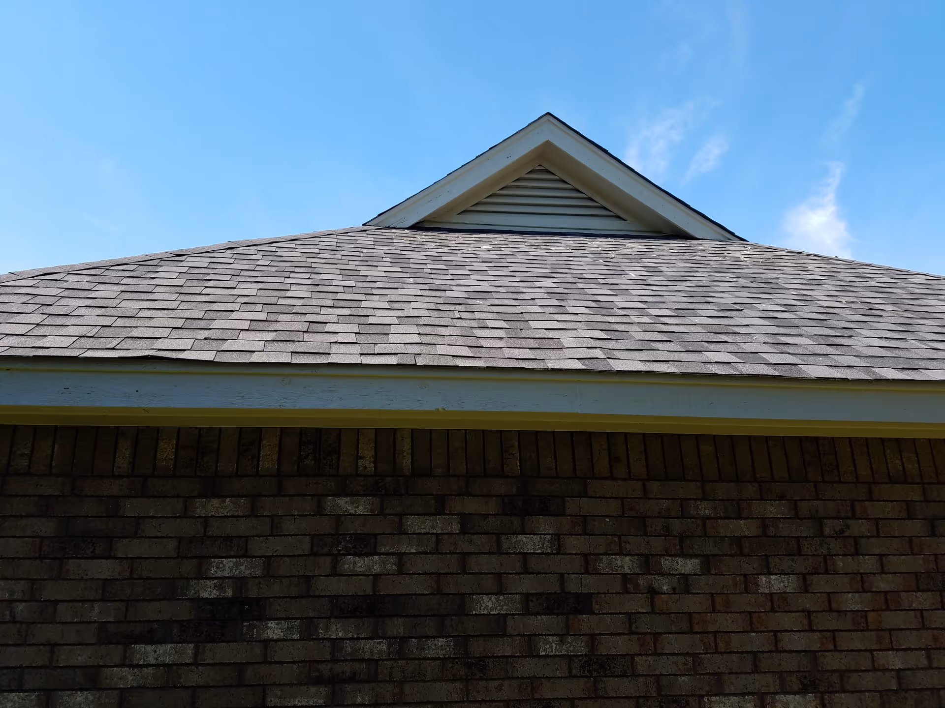 Sloped shingled roof above a brick wall with blue sky.