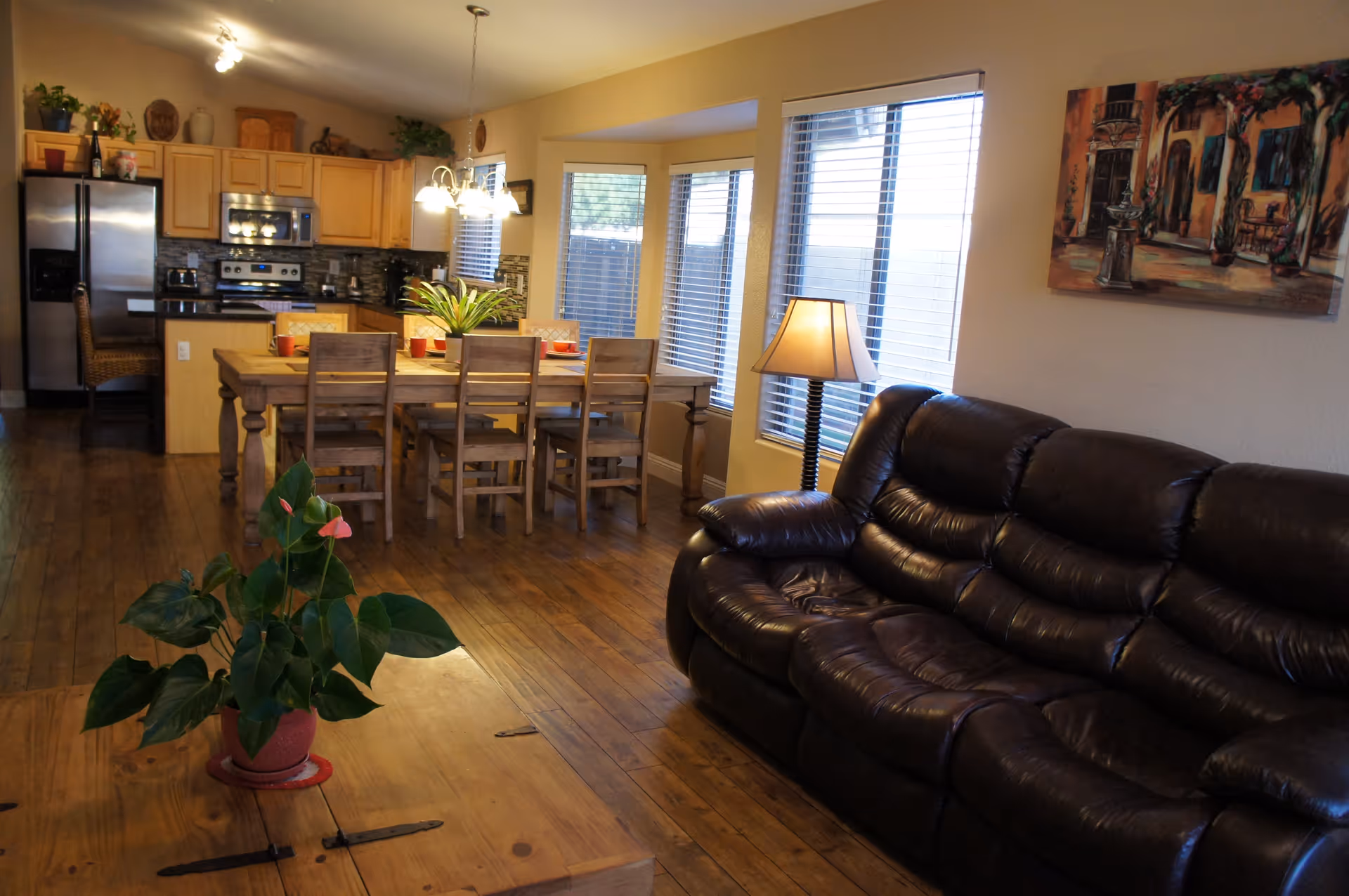 A cozy living and dining area with a dark brown leather sofa on the right, a wooden coffee table with a potted plant in the foreground, and a wooden dining table with six chairs in the middle. The kitchen area with stainless steel appliances is visible in the background. The room has wooden flooring, beige walls, several windows with blinds, a floor lamp, and a colorful painting on the wall.