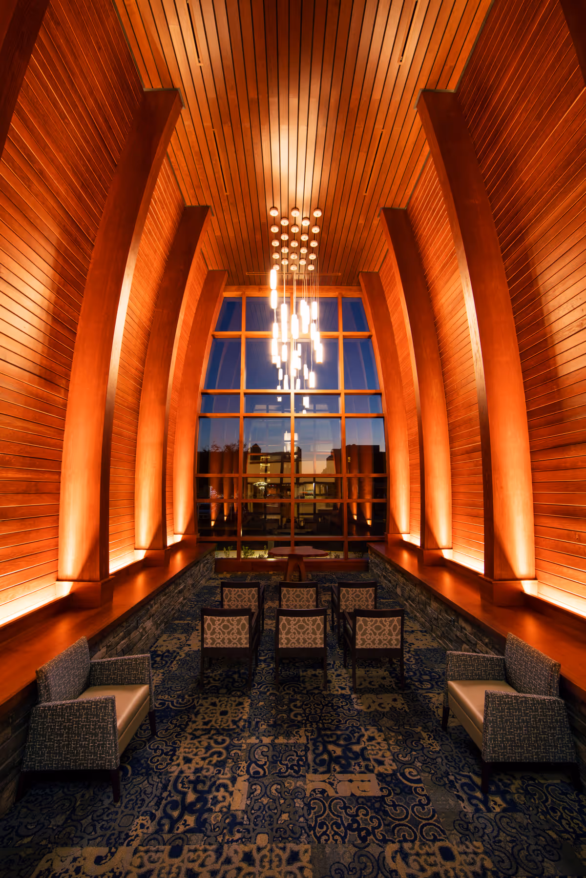 Warm wooden arched walls frame a chandeliered lounge with rows of chairs and a blue rug.