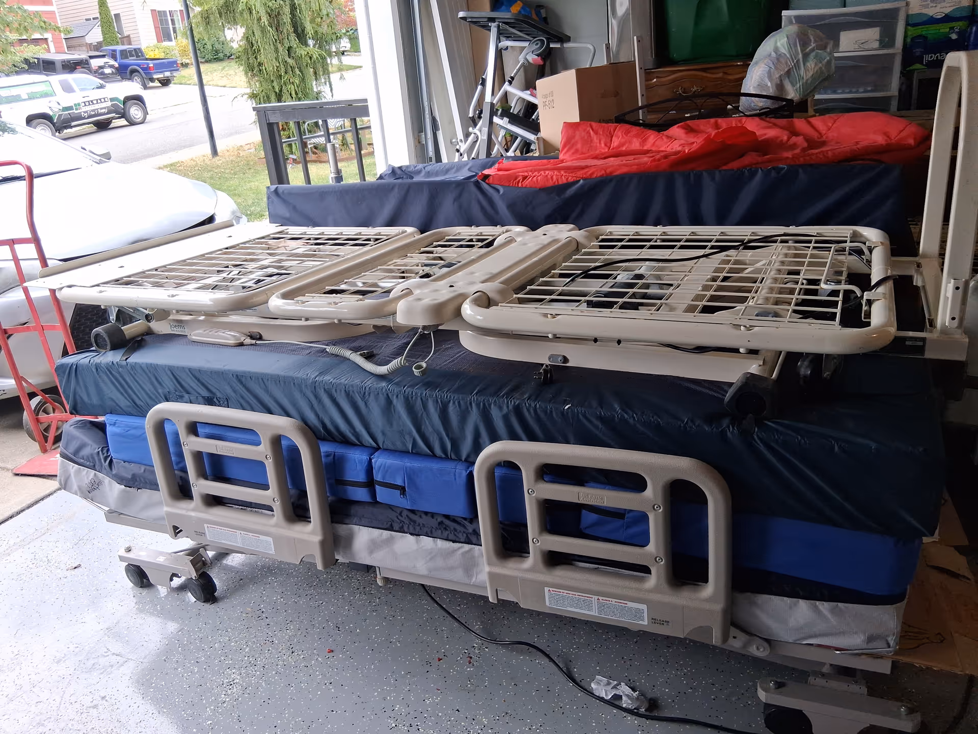 A hospital-style adjustable bed with rails sits in a cluttered garage storage space.