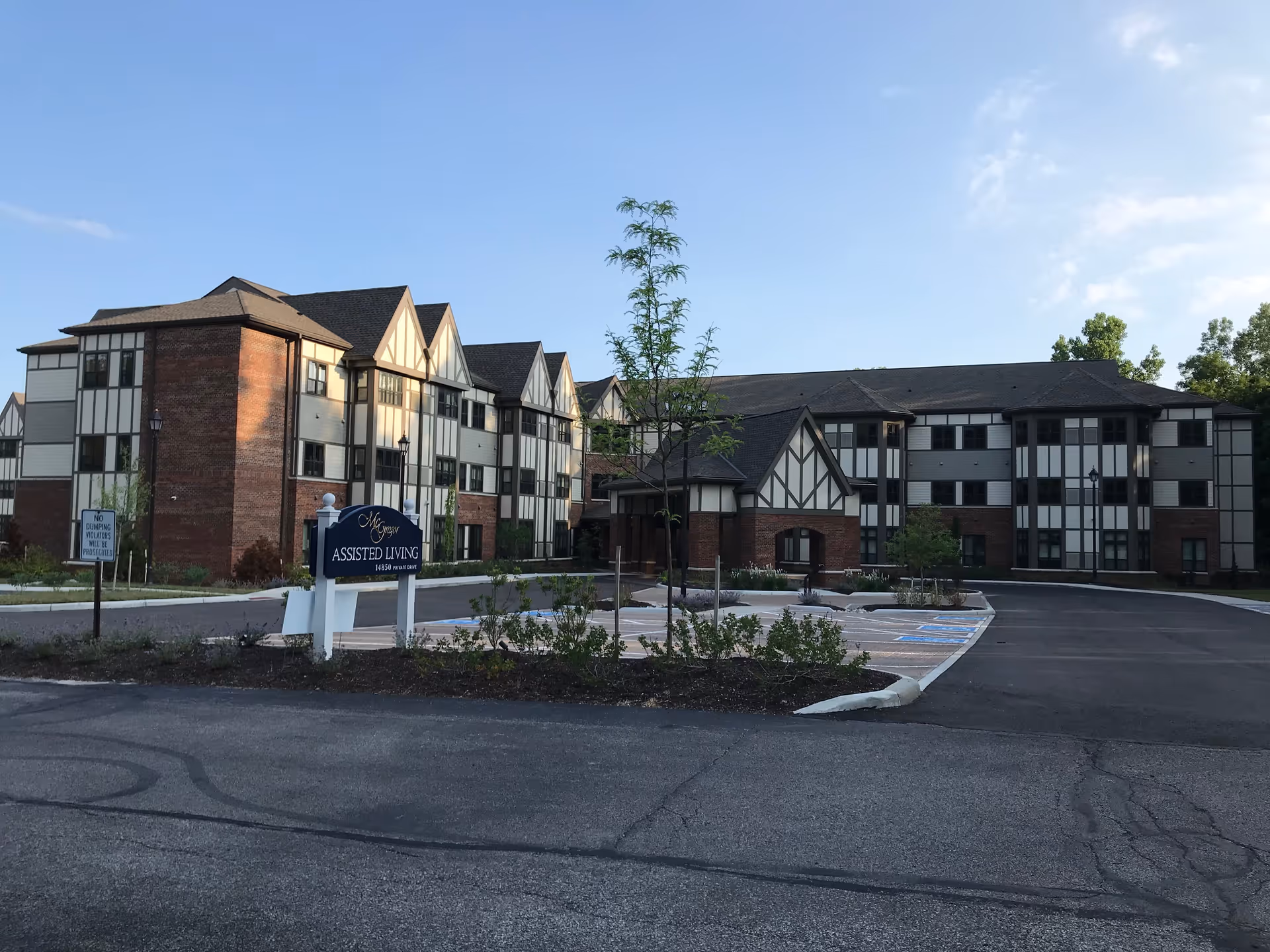 Exterior view of McGregor Assisted Living facility, a multi-story building with a Tudor-style design featuring brick and white paneling. The building is surrounded by a parking lot with marked spaces and some landscaping including small trees and shrubs. A sign in front reads 'McGregor Assisted Living'.