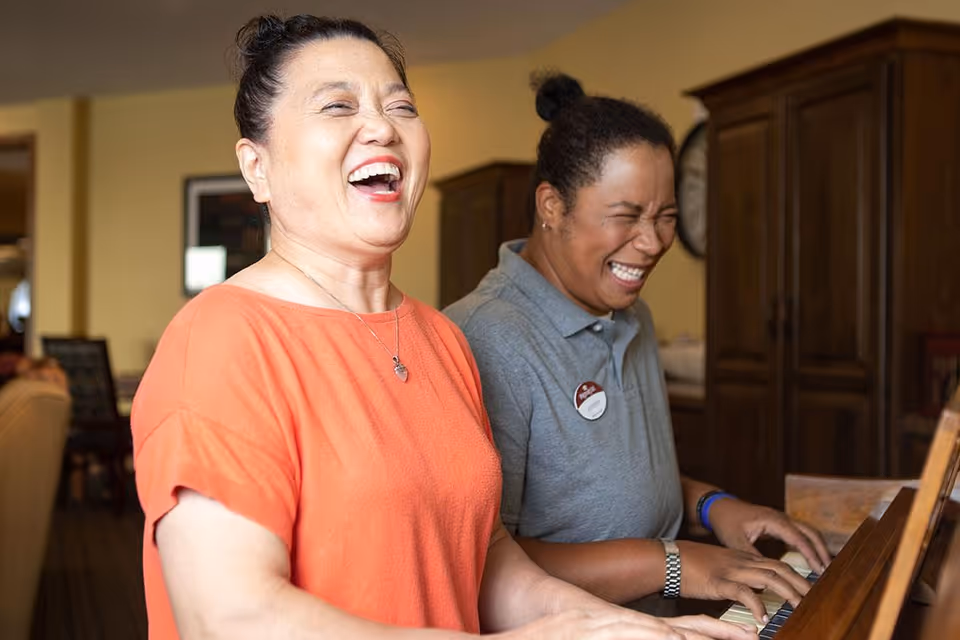 Two women laughing joyfully while playing a piano together in a warmly lit room with wooden furniture in the background.