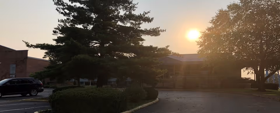 Sunlit exterior of a senior living facility with trees, a driveway, and a parked car at sunset.
