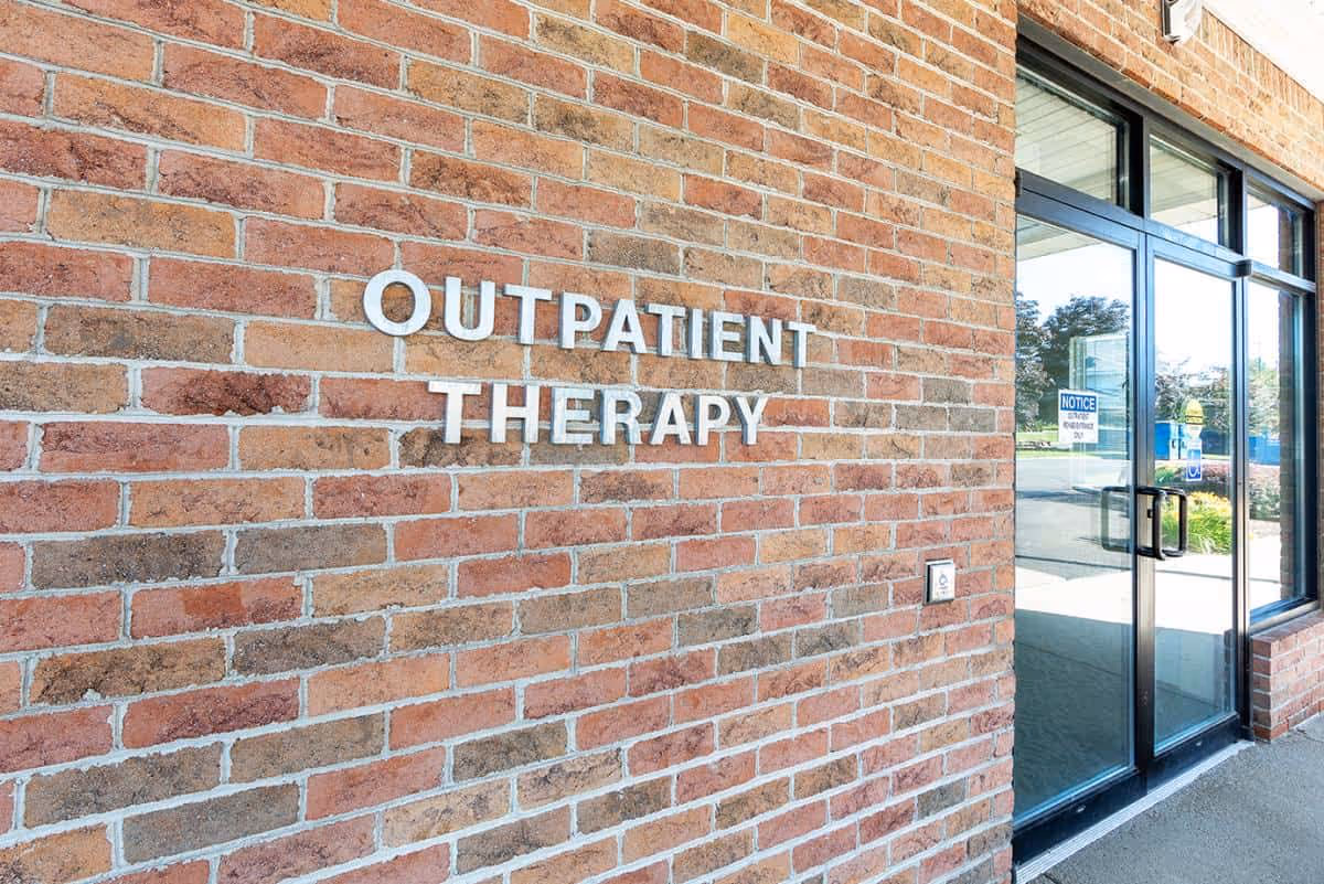 Brick exterior wall with metal letters reading 'OUTPATIENT THERAPY' beside a glass entrance door.