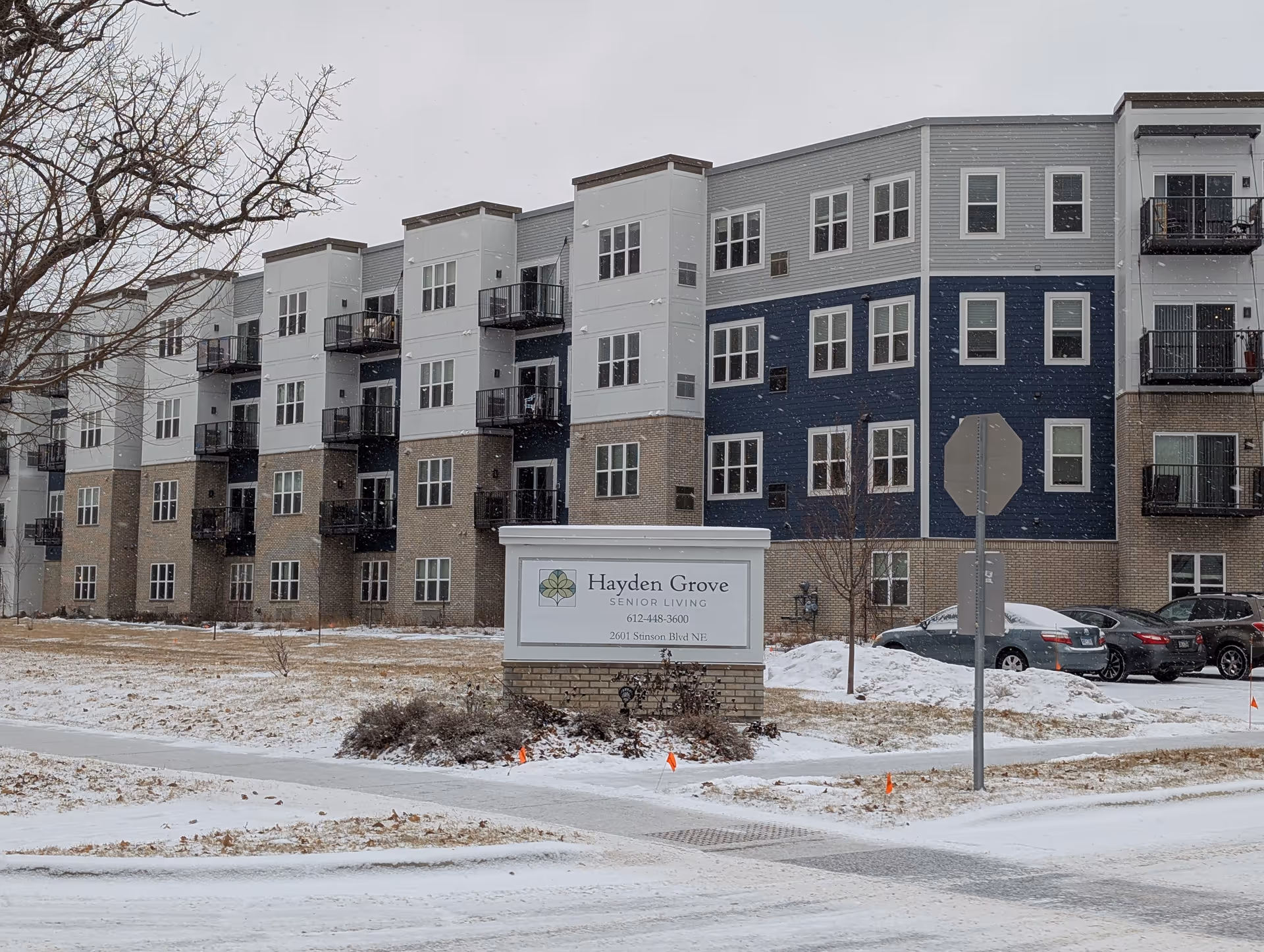 Exterior view of a multi-story senior living facility building with a mix of brick and siding facade, several balconies, parked cars, and a sign in front that reads Hayden Grove Senior Living with contact information. Snow is lightly covering the ground and falling from the sky.