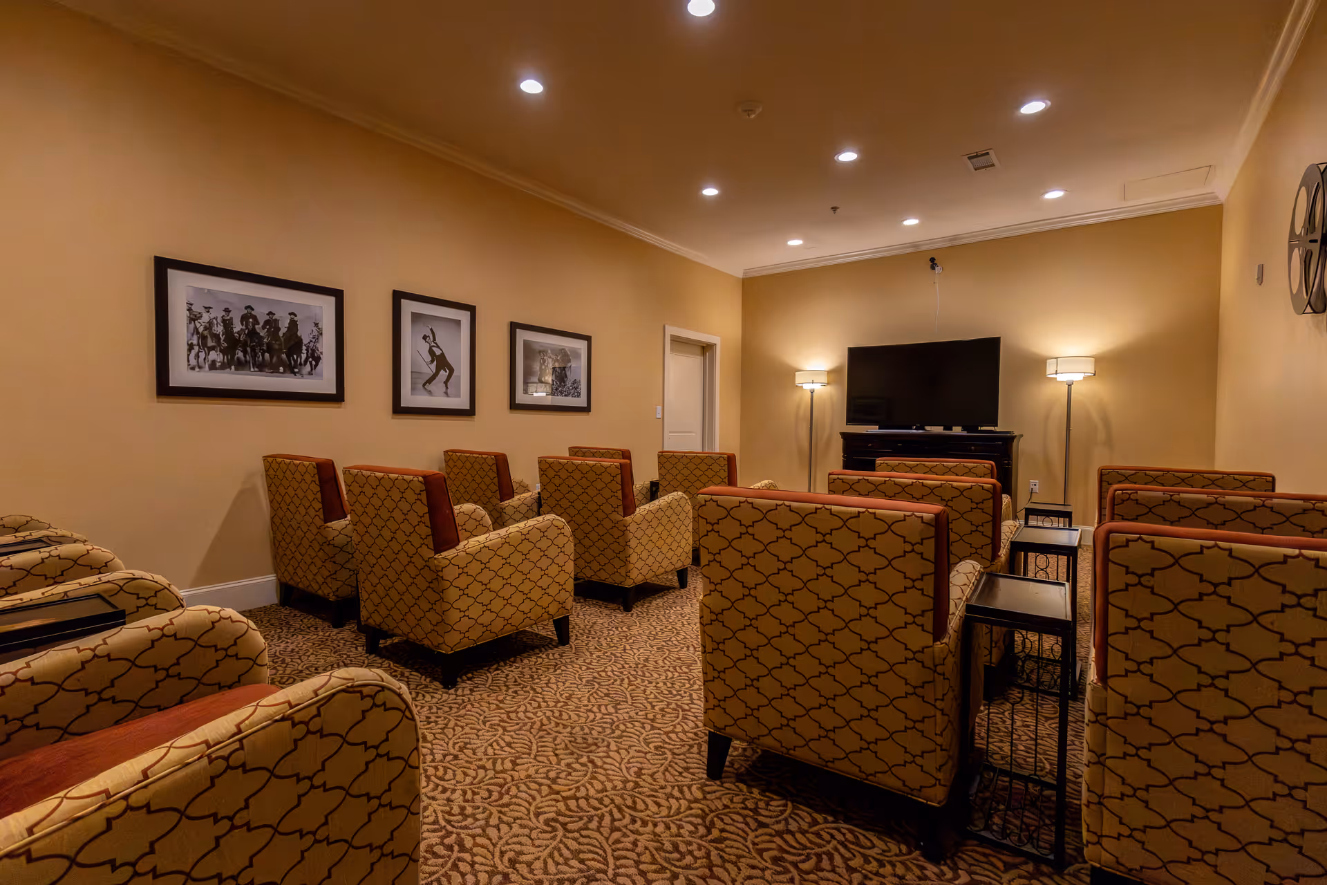 A cozy common room with multiple patterned armchairs arranged in rows facing a flat-screen TV mounted on a stand. The room has warm beige walls, carpeted floor with a floral pattern, three framed black and white photos on one wall, and two floor lamps providing soft lighting.
