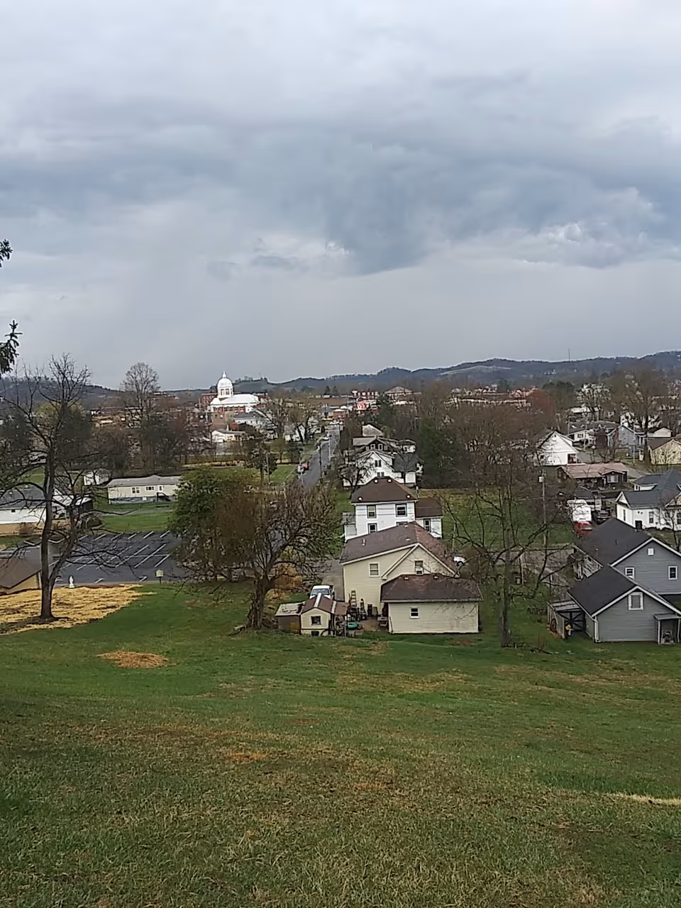 Wide view of a small town with houses on a grassy hill and cloudy sky.