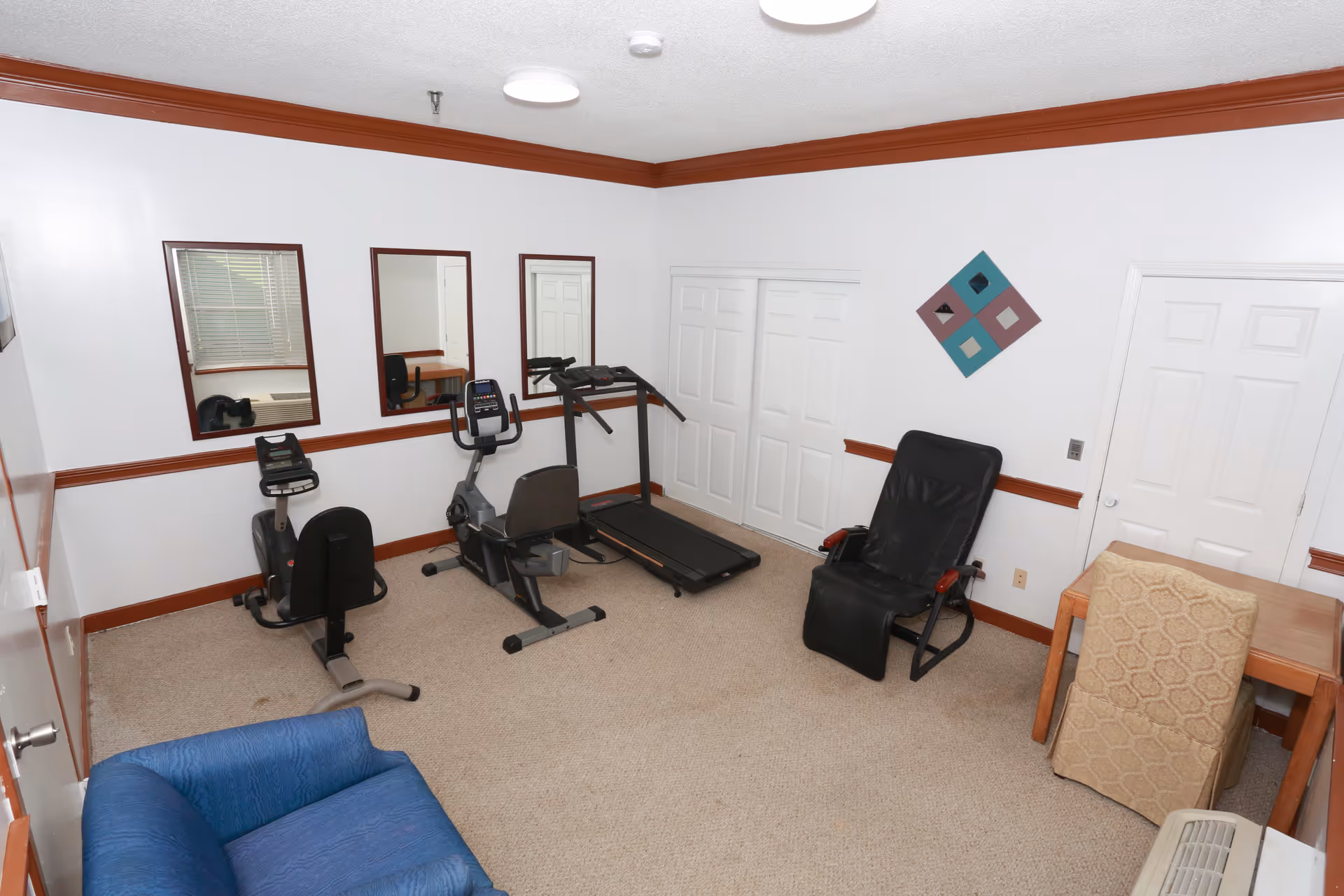 A small exercise room with white walls and brown trim, containing a treadmill, two stationary exercise bikes, a black reclining chair, a blue armchair, a wooden table with a beige upholstered chair, and three rectangular mirrors on one wall.