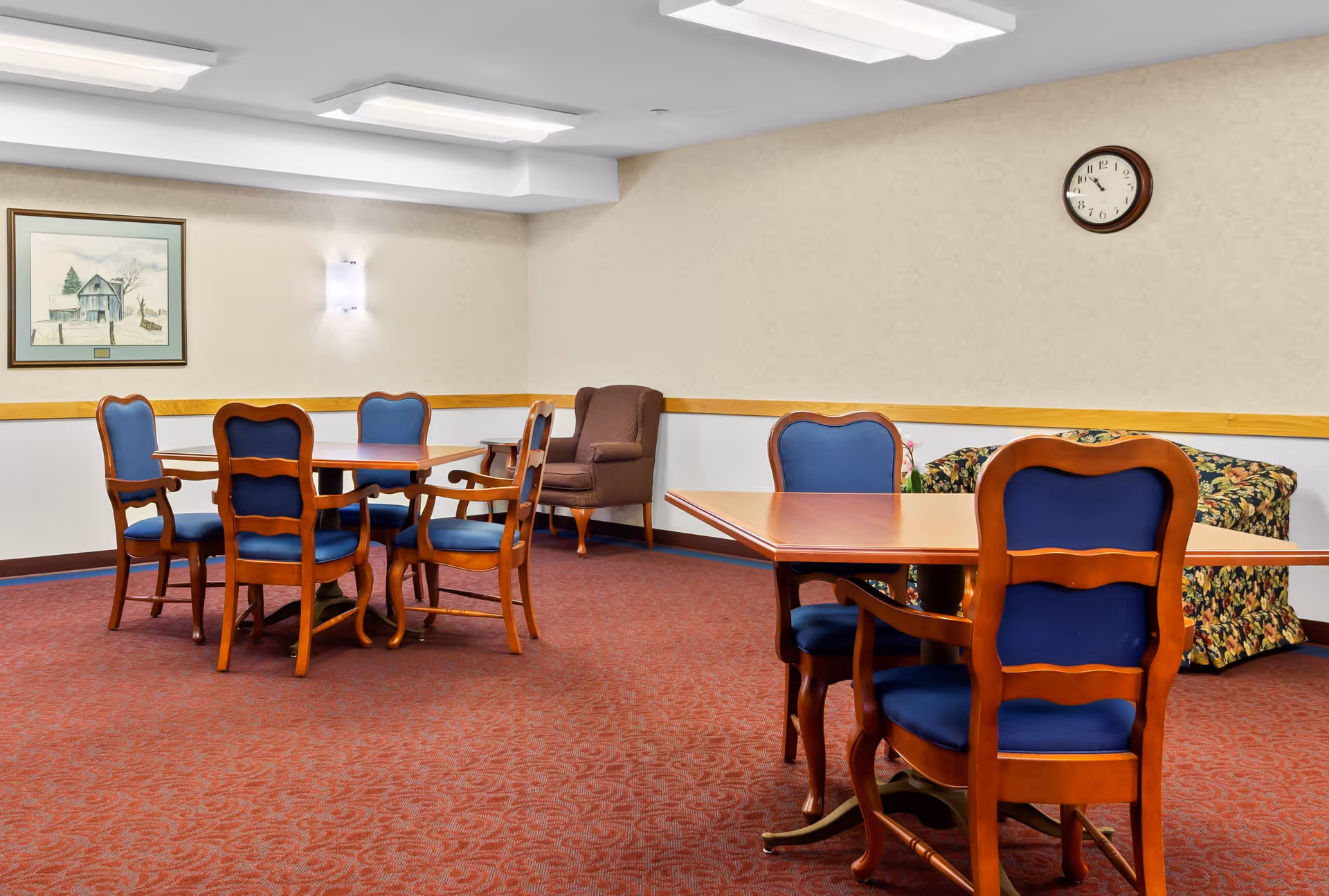 A senior living facility common room with two wooden tables surrounded by blue cushioned chairs. There is a brown armchair and a floral-patterned sofa against the beige walls. A framed picture and a wall clock are also visible on the walls. The floor is covered with red patterned carpet.
