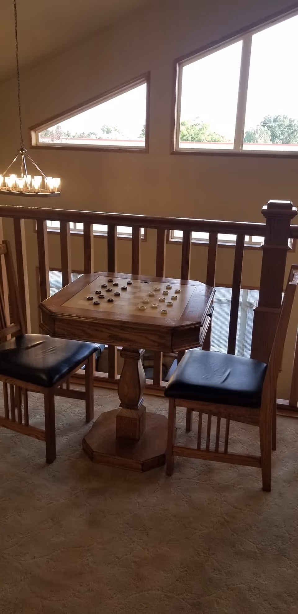 A wooden checkers table with black and white checkers pieces set up, accompanied by two wooden chairs with black cushioned seats, situated in a carpeted area near a wooden railing with large windows in the background letting in natural light.