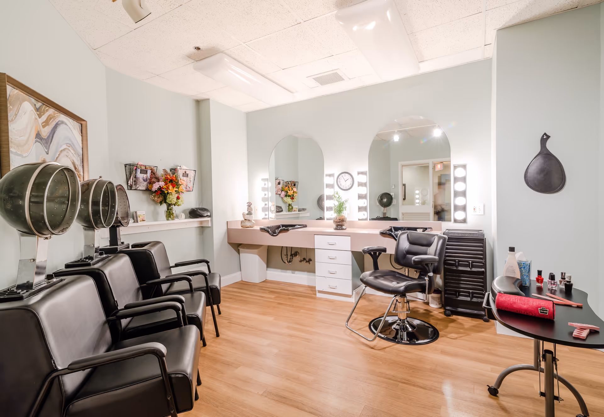 Bright salon interior with multiple styling chairs and hooded dryers, lighted vanity mirrors, and a small manicure station.