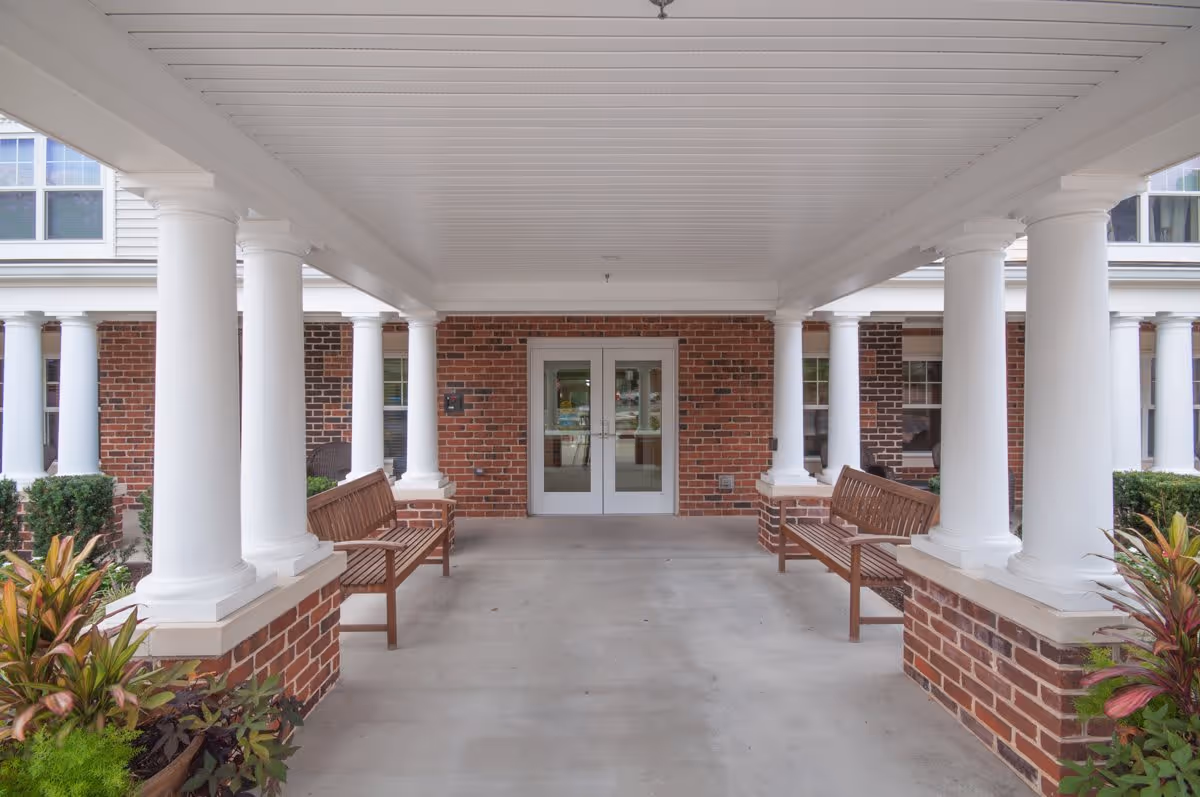 Covered entrance area of a senior living facility with white columns, brick walls, two wooden benches on either side, and double glass doors at the end.