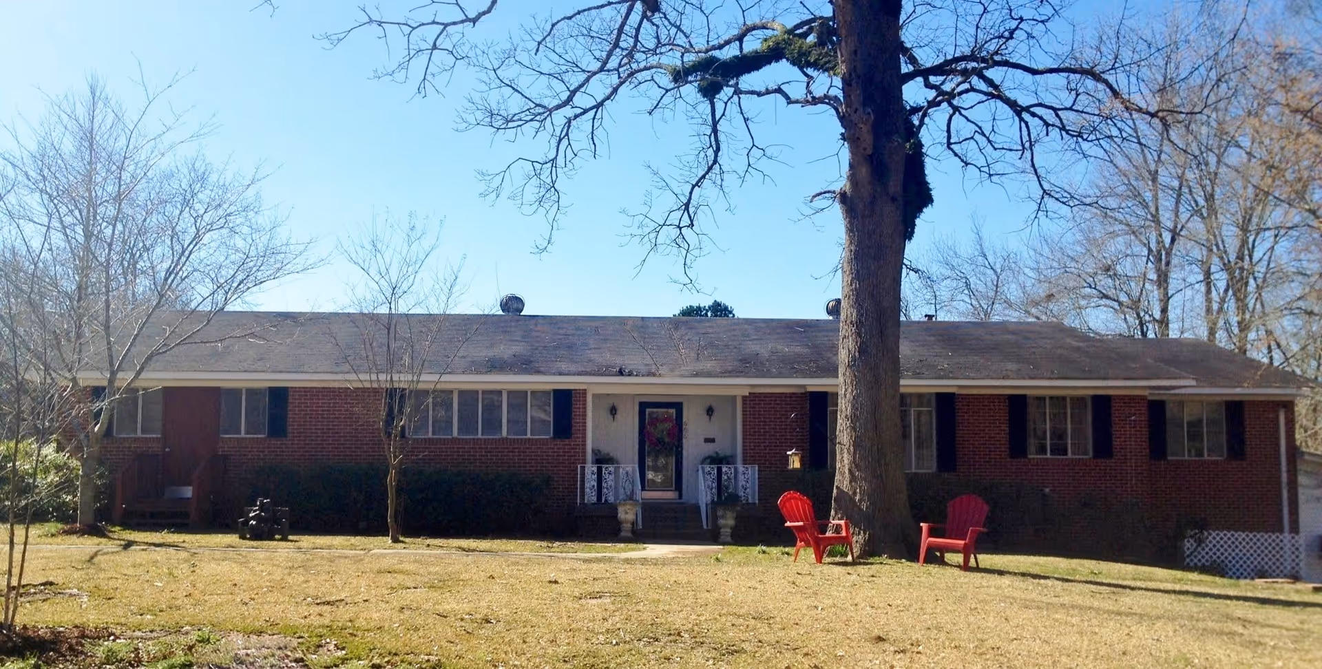 Single-story brick building with a dark shingled roof, several windows with black shutters, and a central entrance with white railings and potted plants. In front of the building is a large tree and two red outdoor chairs on a grassy lawn. The sky is clear and blue.