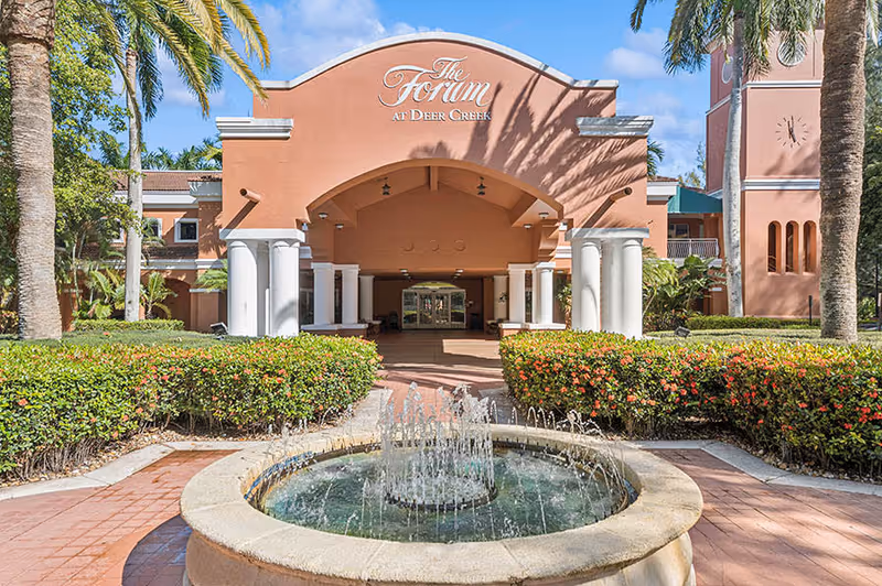 Front exterior view of The Forum at Deer Creek building with a circular water fountain in the foreground, surrounded by green bushes and palm trees under a blue sky.