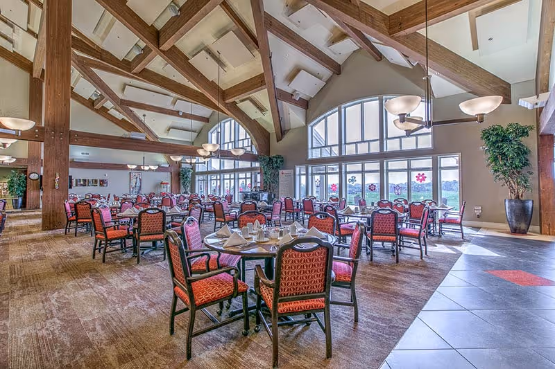 A spacious dining hall with red upholstered chairs around round tables under wooden beams.
