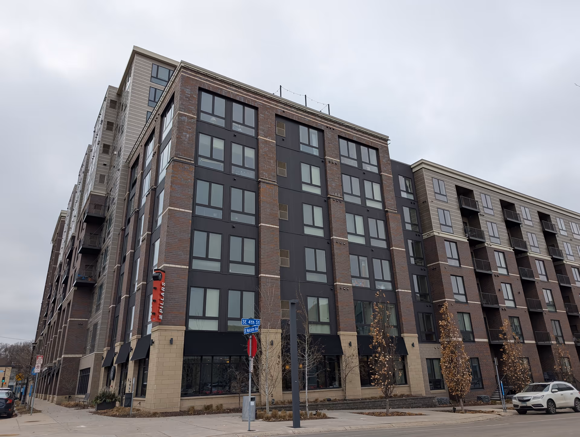 Exterior view of a multi-story brick-and-glass apartment building on a street corner with a vertical red sign reading "Pillars" under a cloudy sky.