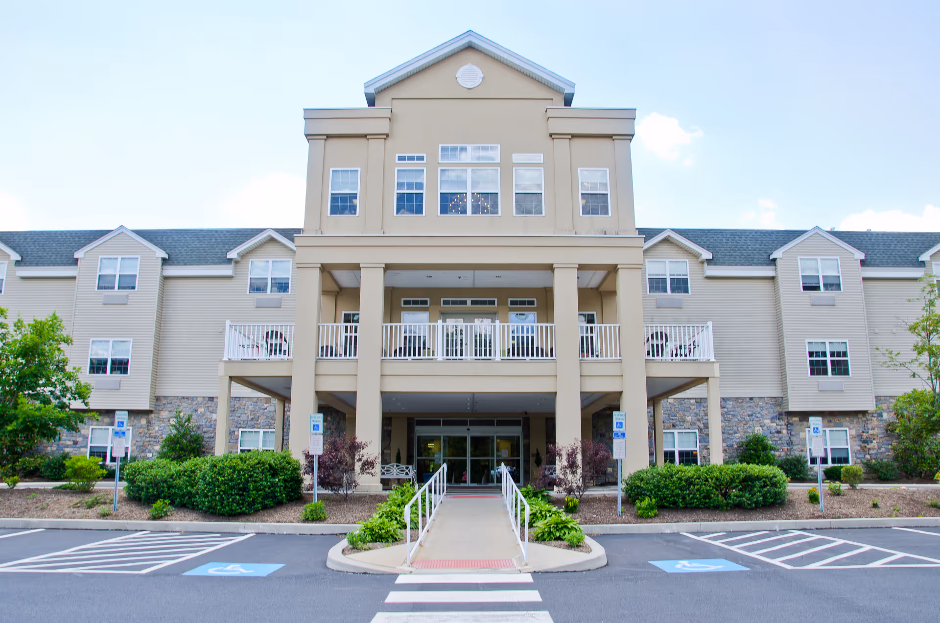 Front exterior view of a multi-story senior living facility building with beige siding and stone accents. The entrance features a covered porch with columns, a ramp, and a walkway leading to glass doors. There are multiple windows and balconies on the upper floors, surrounded by landscaped bushes and trees. Handicap parking spaces are visible in the foreground.
