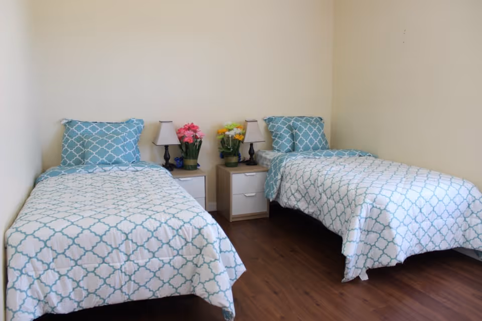 A simple bedroom with two twin beds placed parallel to each other against cream-colored walls. Each bed has a matching blue and white patterned comforter and pillow. Between the beds are two small nightstands with table lamps and colorful flower arrangements. The floor is wooden.