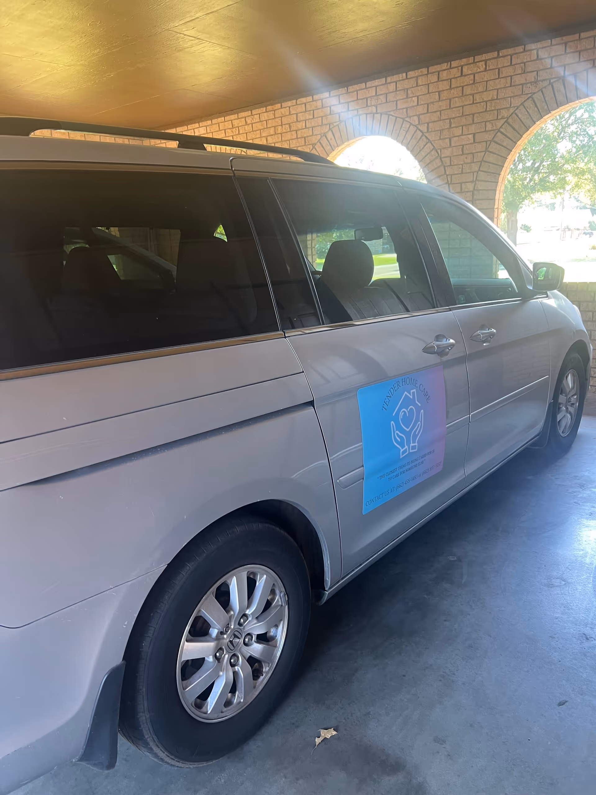 Silver passenger van with Tender Home Care logo parked under a brick arch carport.