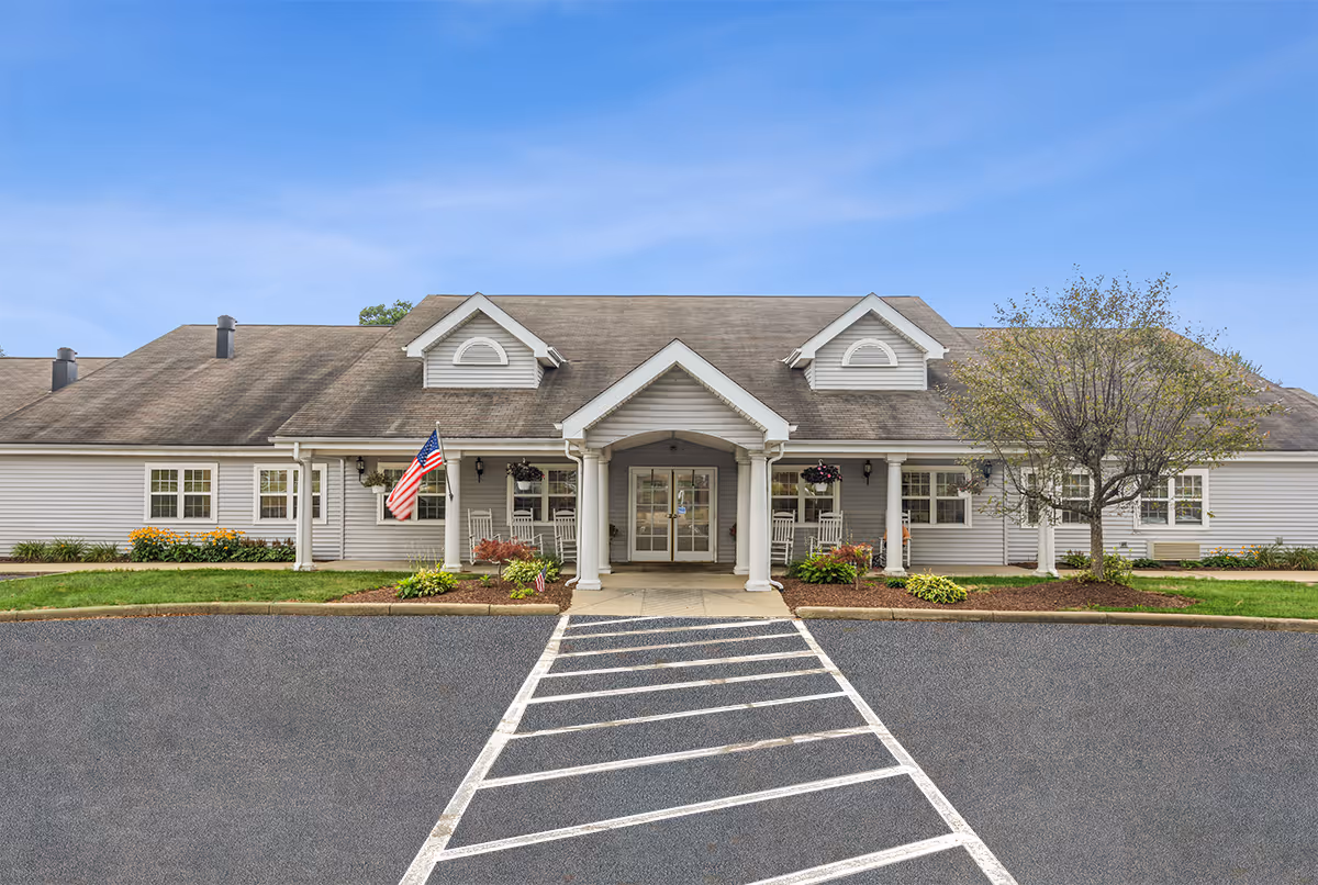 Single-story senior living facility with covered entrance, white columns, flag, and manicured landscaping.