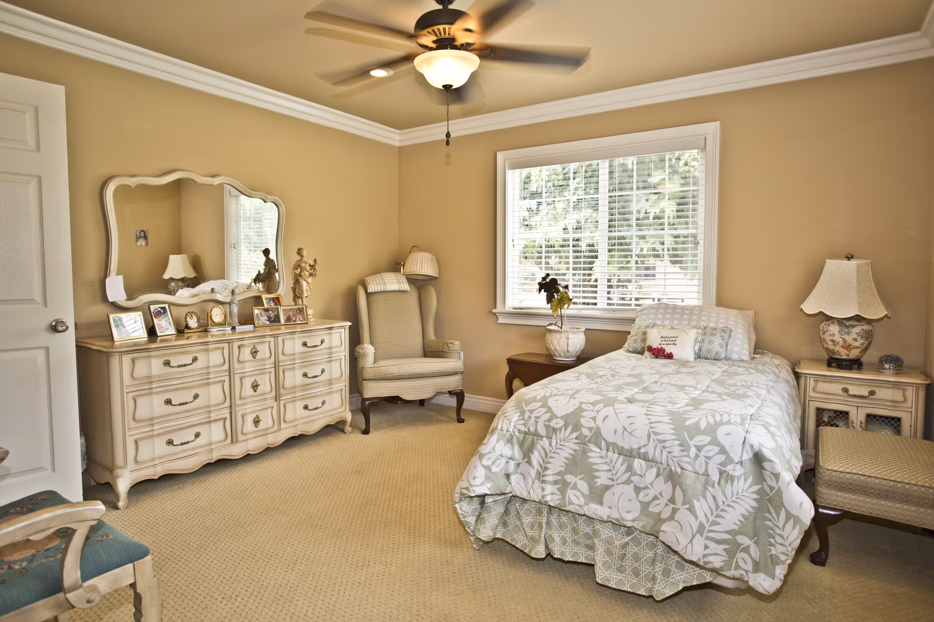 A cozy bedroom with beige walls and carpet. The room features a single bed with a green and white leaf-patterned comforter and a decorative pillow. Next to the bed is a wooden nightstand with a floral lamp and a small purple decoration. A cushioned armchair with a plaid blanket draped over the back sits near a window with white blinds. Across from the bed is a cream-colored dresser with ornate handles, topped with framed photos, a clock, and decorative statues. A large mirror hangs above the dresser. The ceiling has crown molding and a ceiling fan with a light fixture.