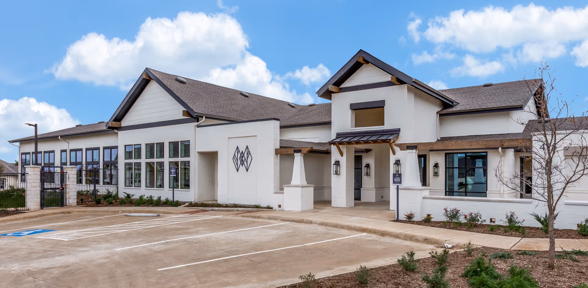 Exterior view of a modern senior living facility building with white walls, large windows, and a sloped roof under a partly cloudy sky. There is a paved parking area with marked spaces and some landscaping with small plants and a tree near the entrance.