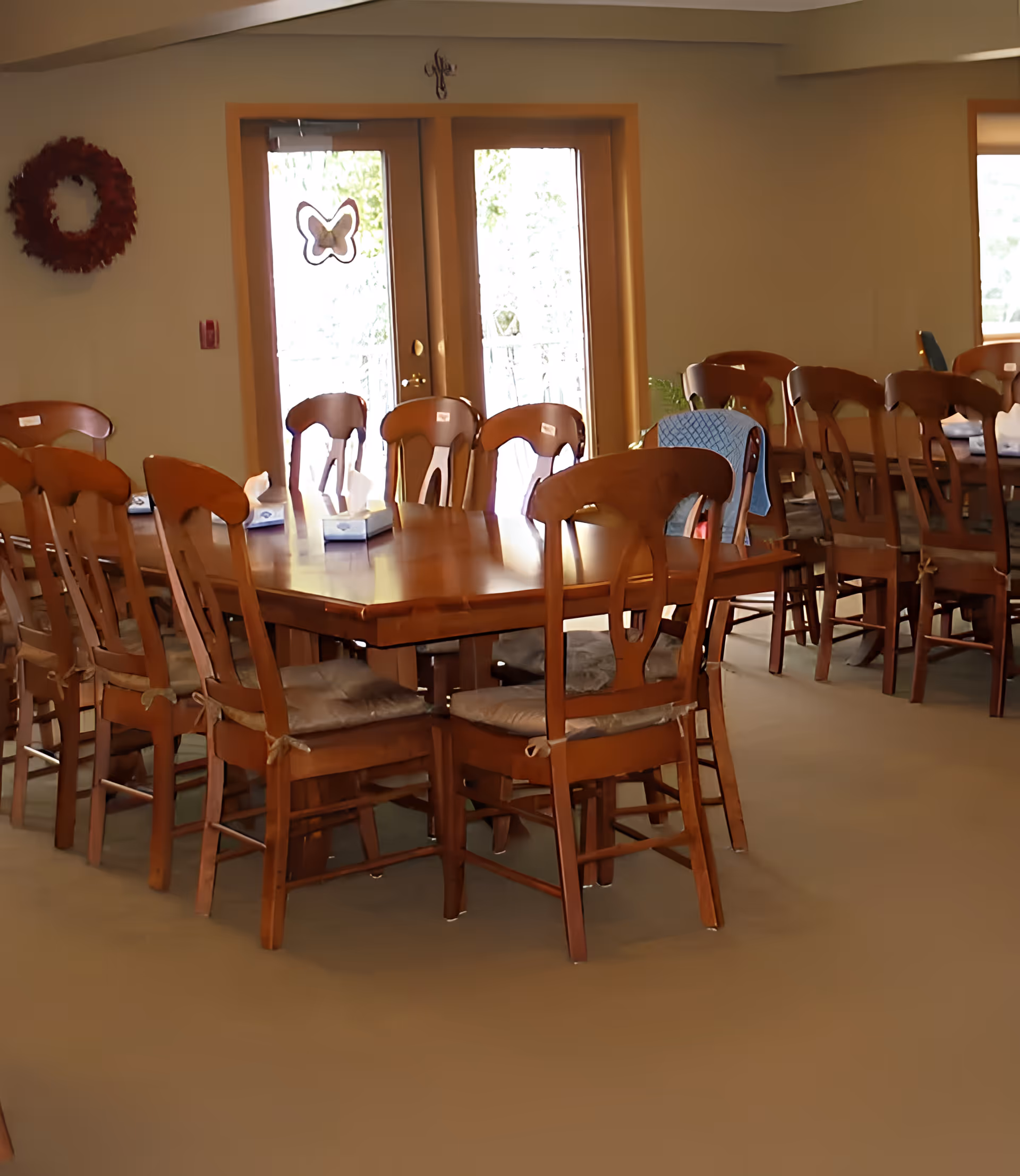 A dining room with multiple wooden tables and chairs arranged neatly. The room has beige walls, a wreath decoration on one wall, and double glass doors with a butterfly decoration leading outside. Tissue boxes are placed on the tables.