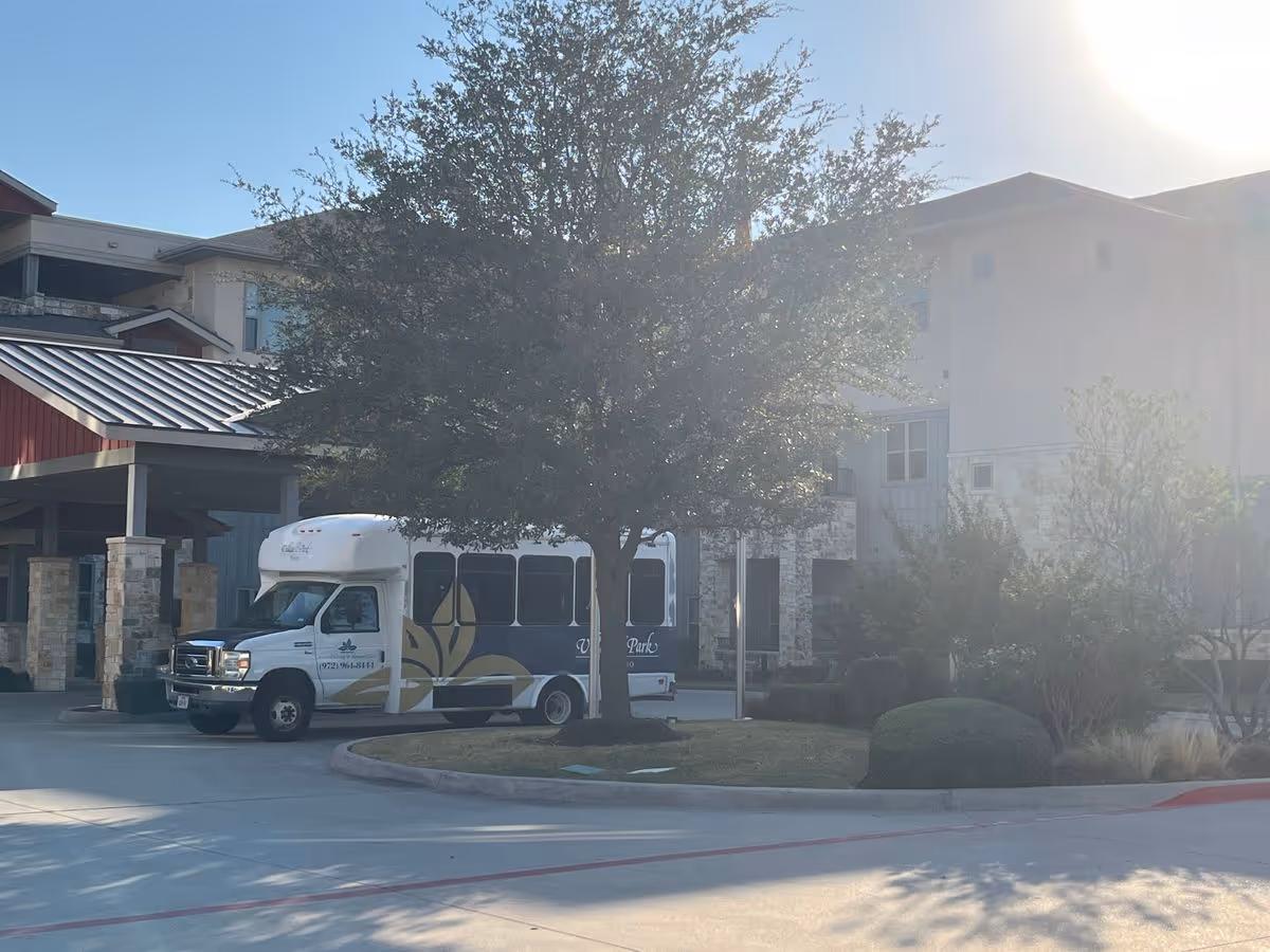 Shuttle bus parked under a tree in front of the entrance to a multi-story senior living building.