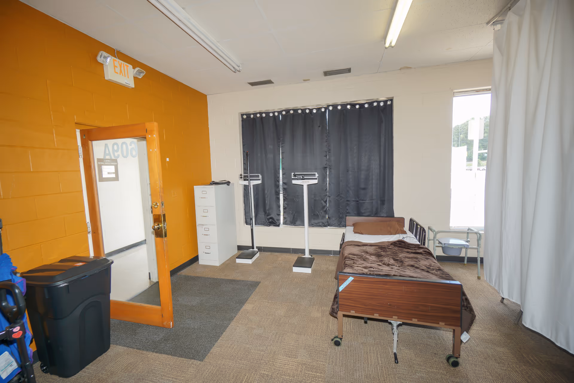 Interior room with a hospital bed, scales, a cabinet, and orange wall near EXIT sign.