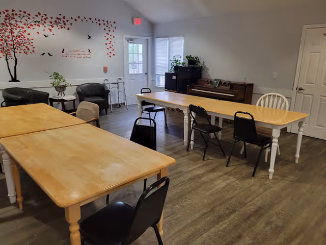 A senior living common room with two long wooden tables and chairs, a piano, lounge chairs, and a tree wall decal.