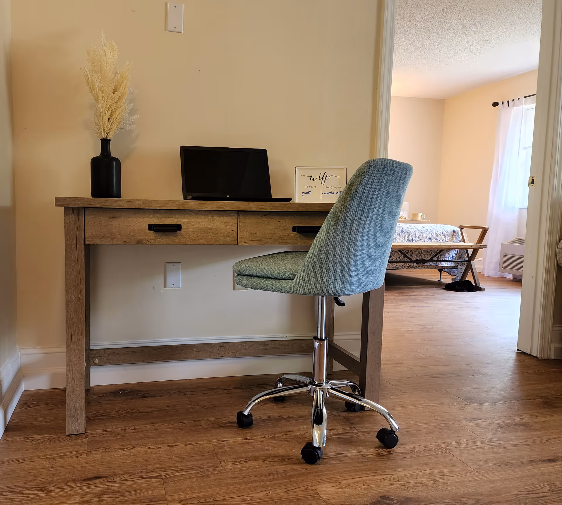 A wooden desk with two drawers against a beige wall, featuring a black vase with dried flowers, a closed laptop, and a small sign with WiFi information. In front of the desk is a light blue office chair with wheels. In the background, an adjacent room is visible with a bed, a bench at the foot of the bed, a window with white curtains, and a heating/cooling unit below the window.