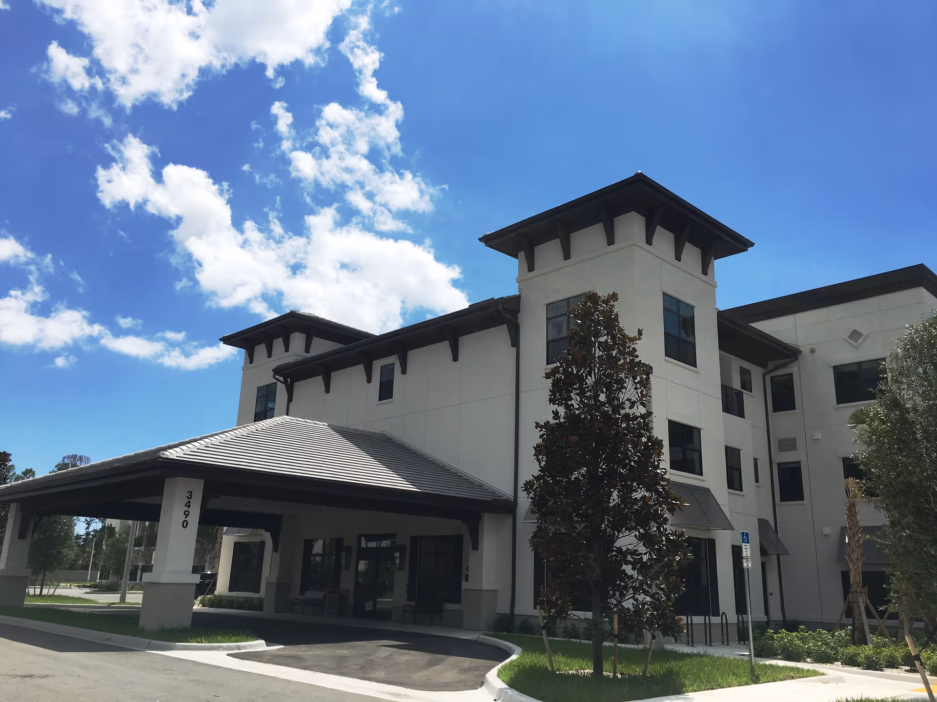 Exterior view of a modern three-story senior living facility building under a blue sky with scattered clouds. The building has a covered entrance with the number 3490 on a pillar, large windows, and landscaped greenery including trees and shrubs.