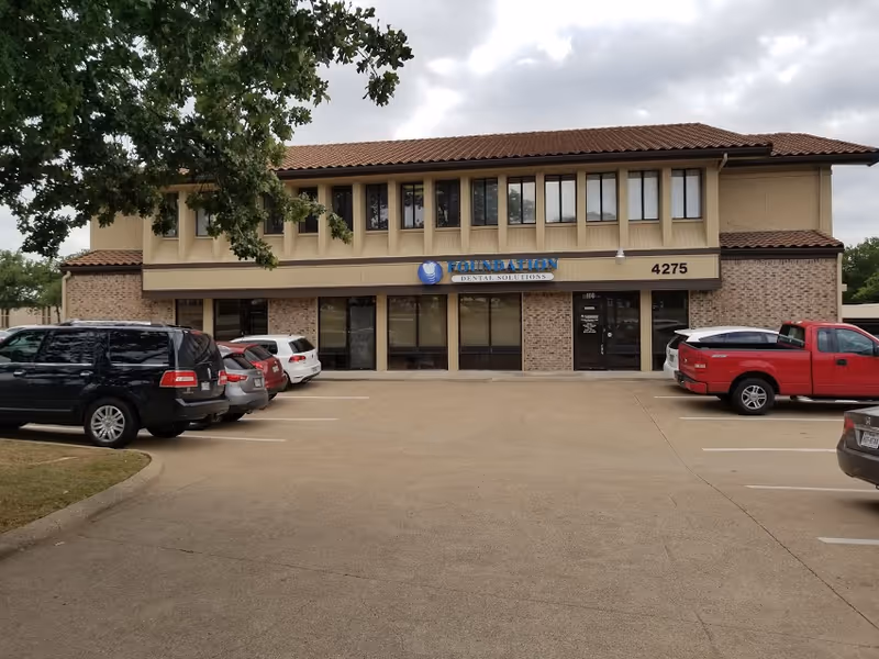 Two-story beige storefront building with large windows and brick facade in a parking lot.