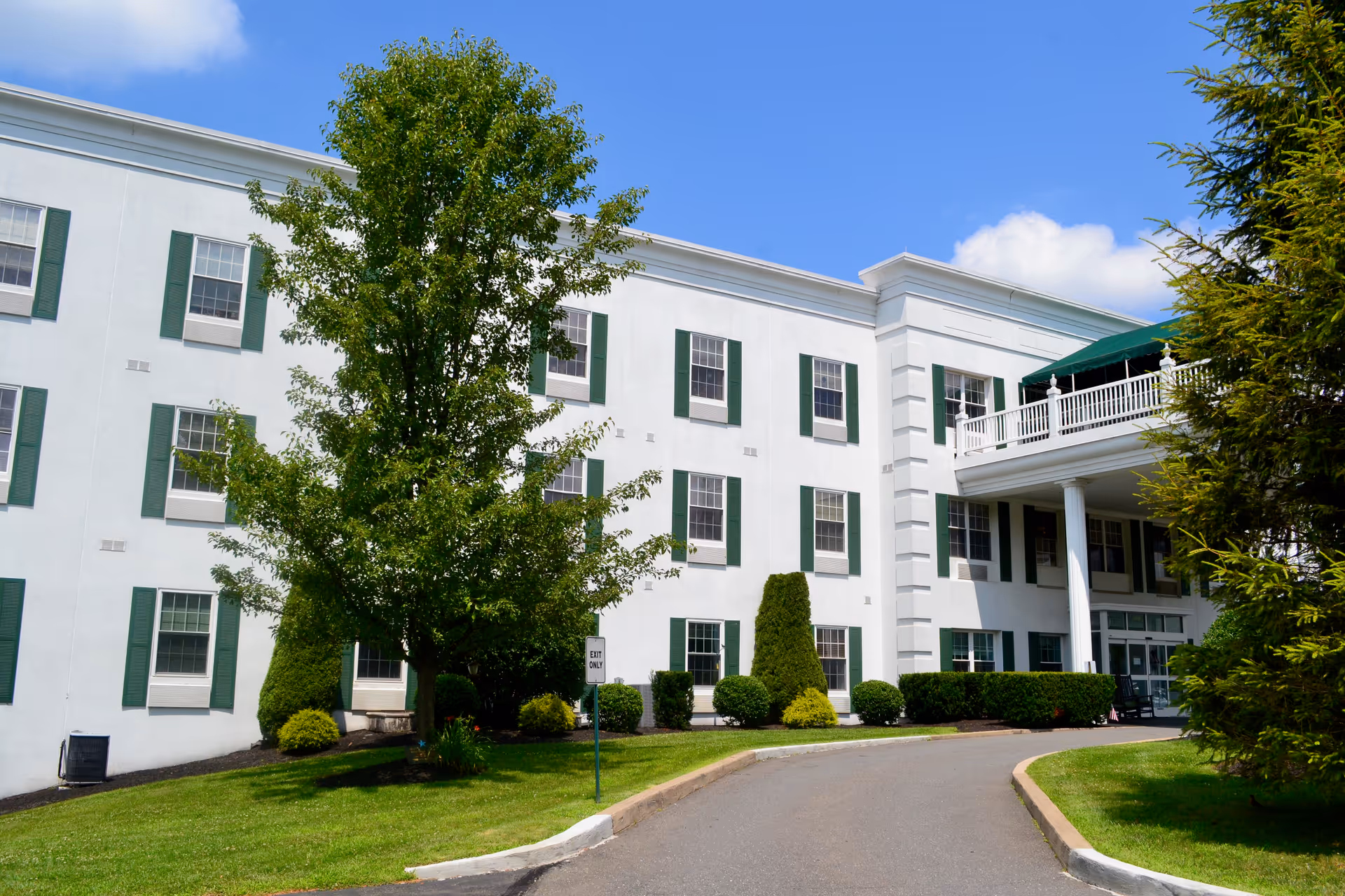 Exterior view of a three-story white senior living facility building with green window shutters, a driveway leading to the entrance, and well-maintained landscaping including trees and bushes under a blue sky with some clouds.