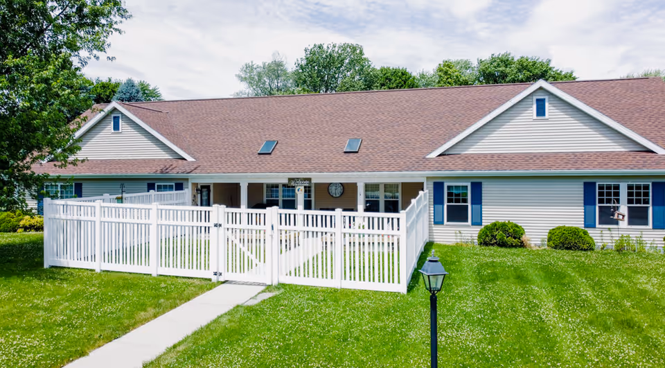 Single-story assisted living building with a white picket fence enclosing the front yard and a walkway to the entrance.