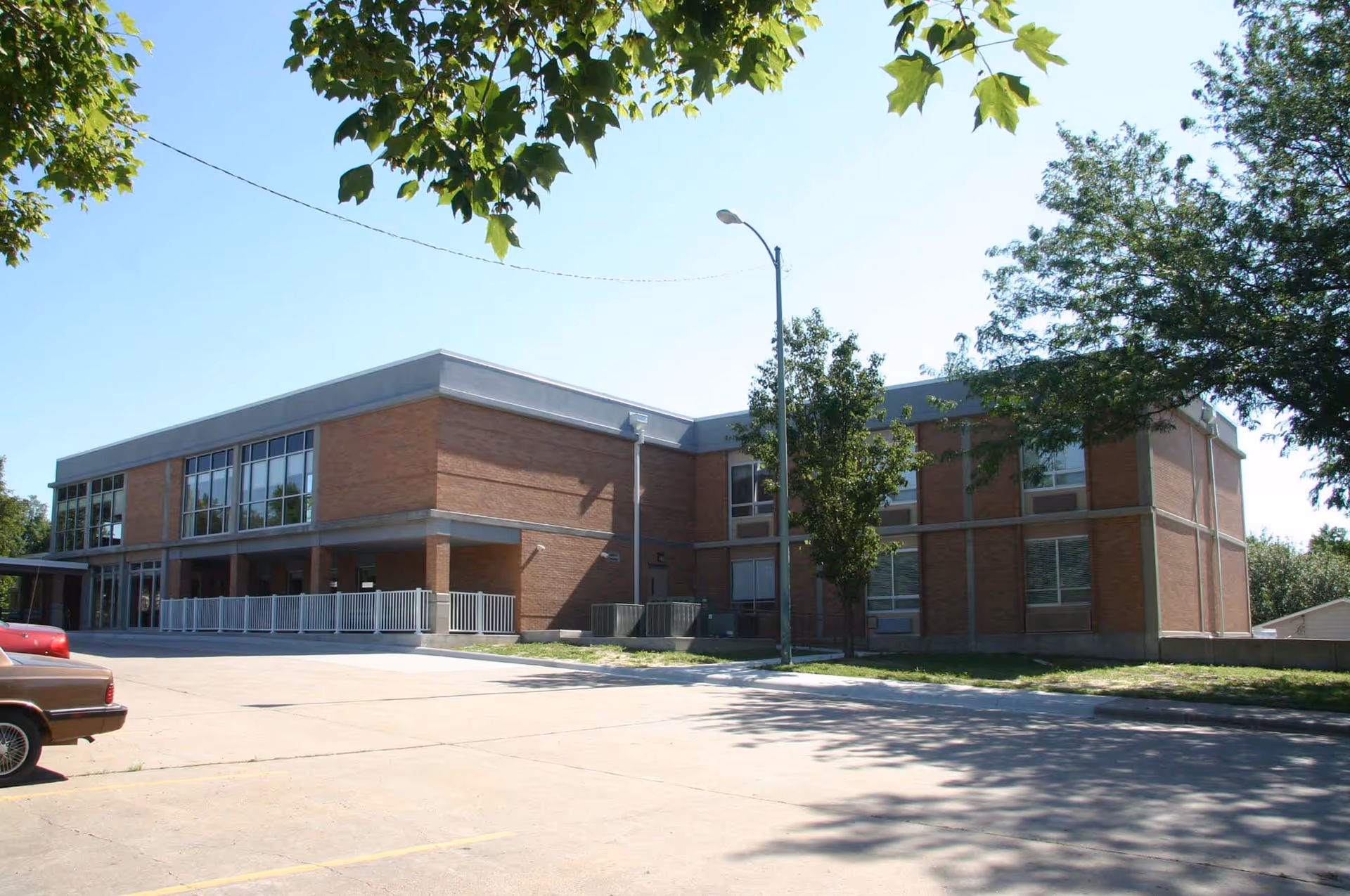 Exterior view of a two-story brick building with large windows and a white railing along the entrance area. There are trees around the building and a parking lot in the foreground with a few parked cars. The sky is clear and blue.