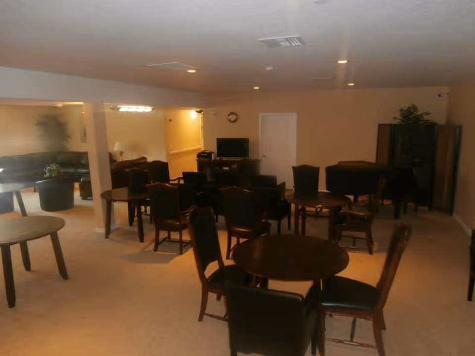 A common area in an assisted living facility with multiple round tables and chairs arranged throughout the room. There is a television on a stand against the far wall, a clock above it, and a piano in the corner. The room has beige walls and carpet, recessed lighting in the ceiling, and some plants for decoration.