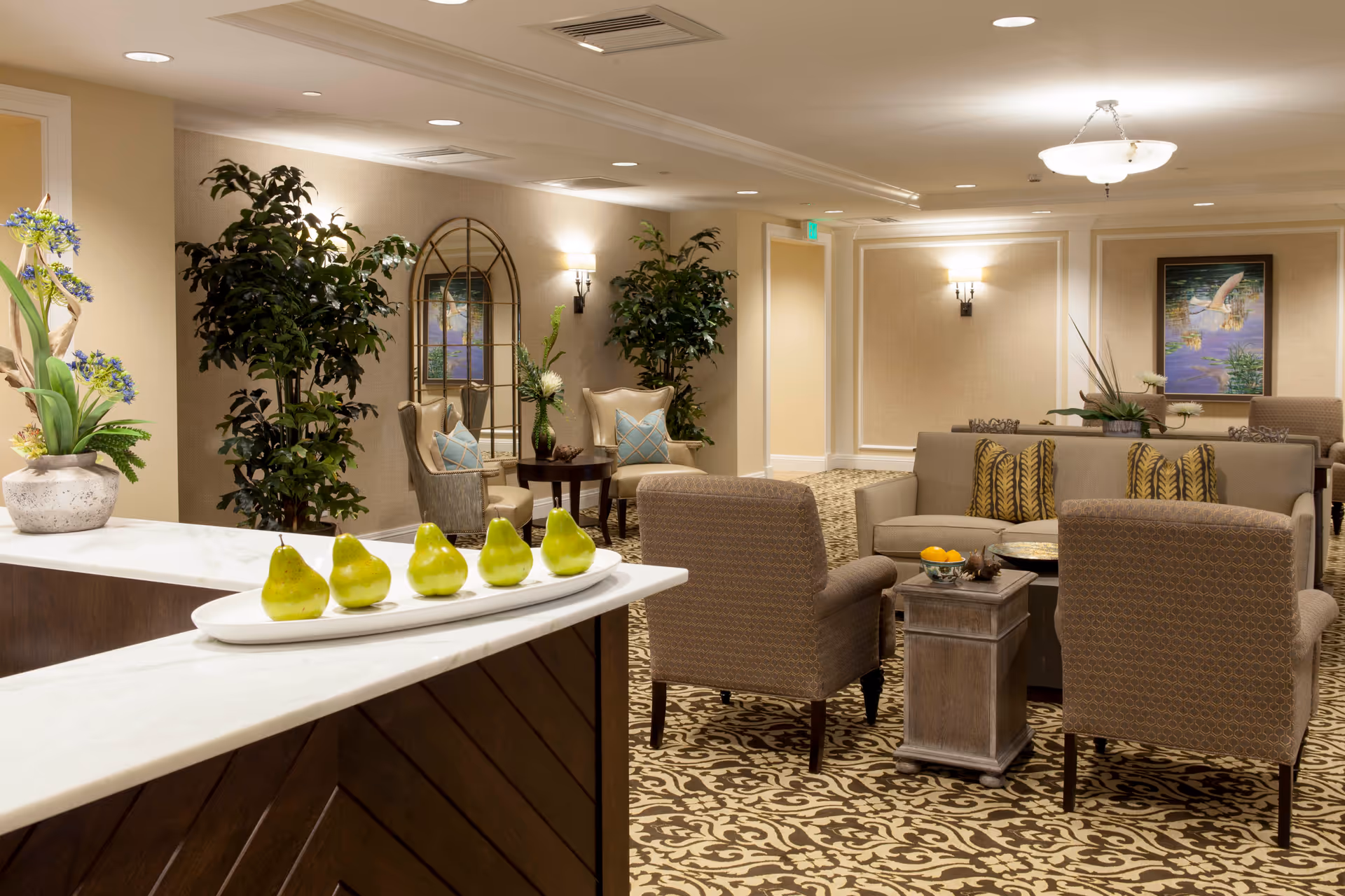 A well-lit senior living facility common area with patterned carpet, beige walls, and comfortable seating including armchairs and sofas with decorative pillows. There are potted plants and floral arrangements, a large mirror on the wall, and framed artwork. A white countertop in the foreground displays a row of green pears on a white platter.