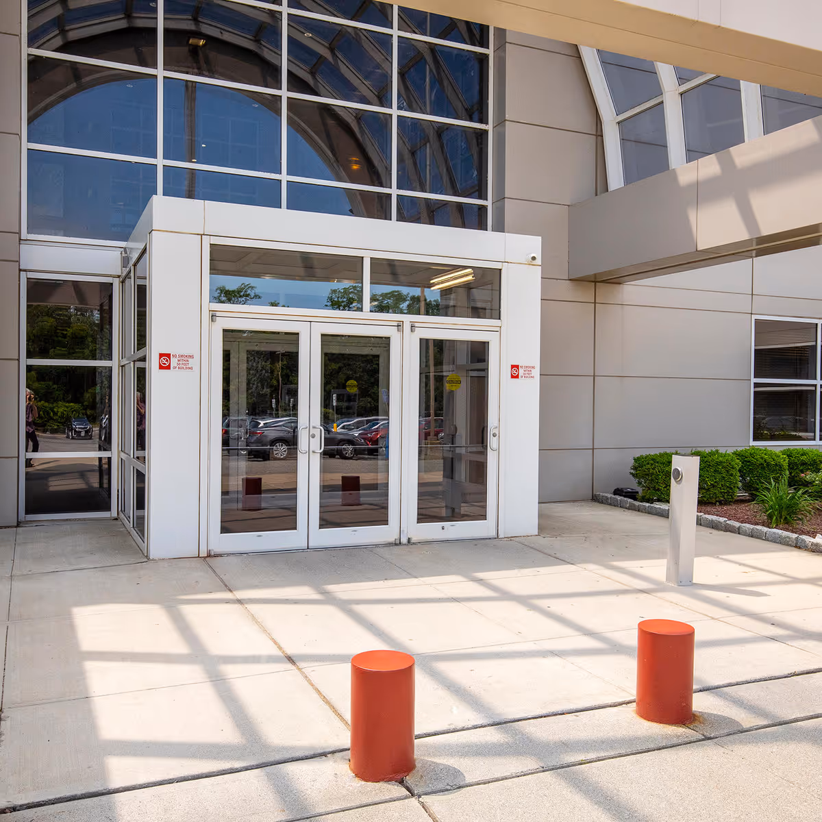 Glass double doors beneath a curved glass atrium entrance, with red bollards and shrubs.