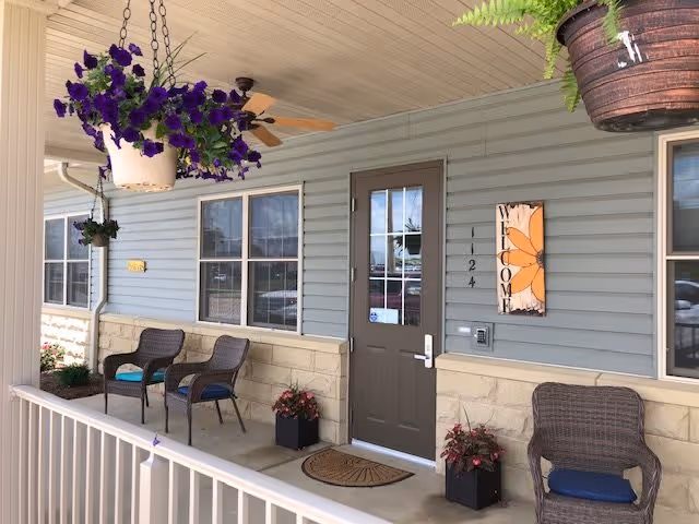 Covered porch area with a brown door labeled 124, two windows, and a welcome sign featuring a large orange flower. The porch has a ceiling fan, hanging flower pots with purple and green plants, three wicker chairs with blue cushions, and two potted plants on the ground near the door. A white railing runs along the edge of the porch.