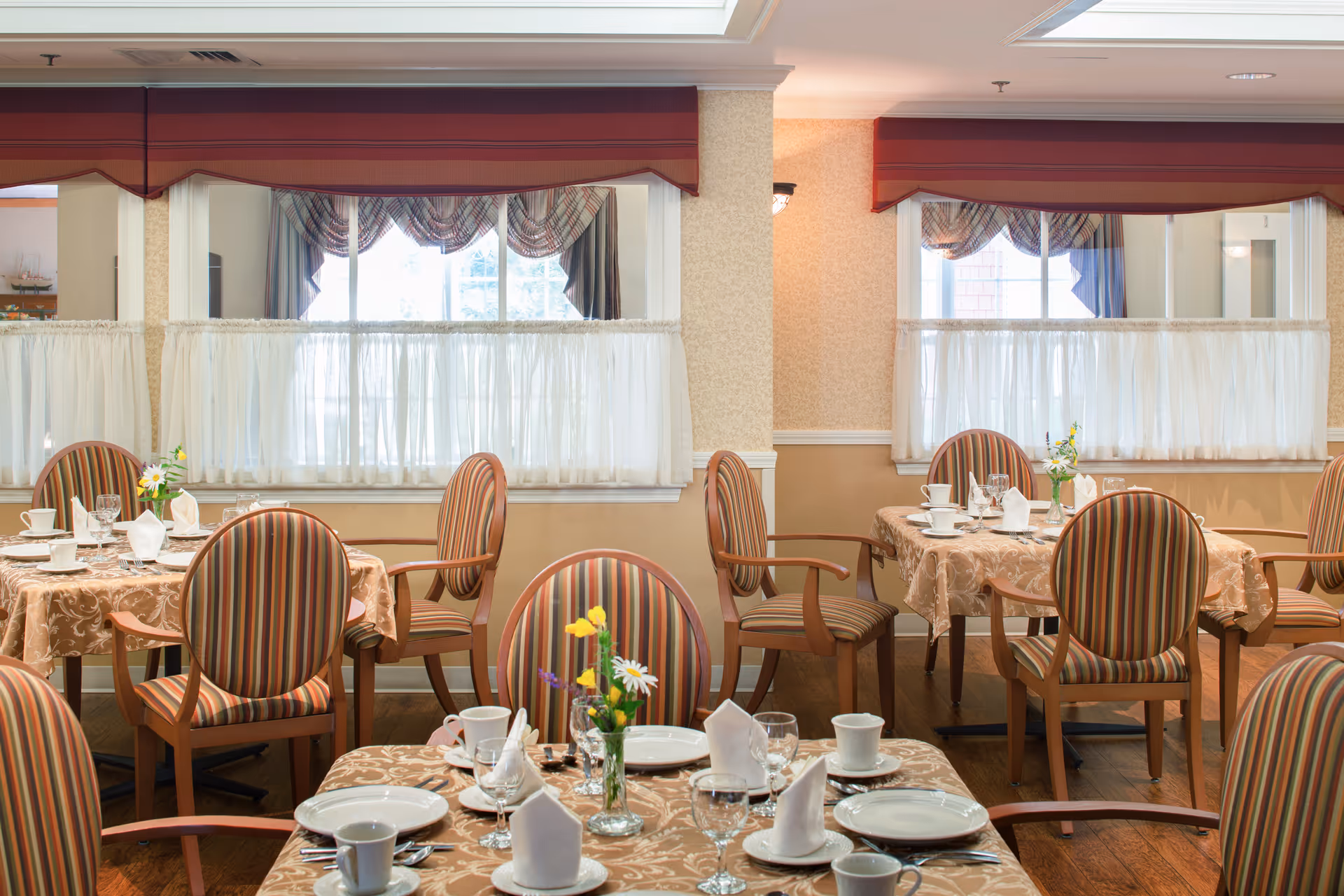 A dining room with tables covered in beige patterned tablecloths, set with white plates, cups, napkins, and glassware. Each table has a small vase with fresh flowers. The chairs have wooden frames and striped upholstery. Large windows with sheer white curtains and valances allow natural light to fill the room.